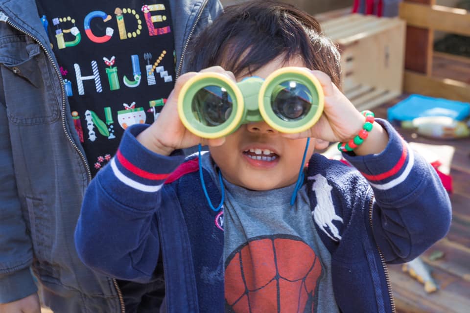 Child holding binoculars up to eyes, smiling, wearing a blue jacket and gray shirt with a basketball graphic.
