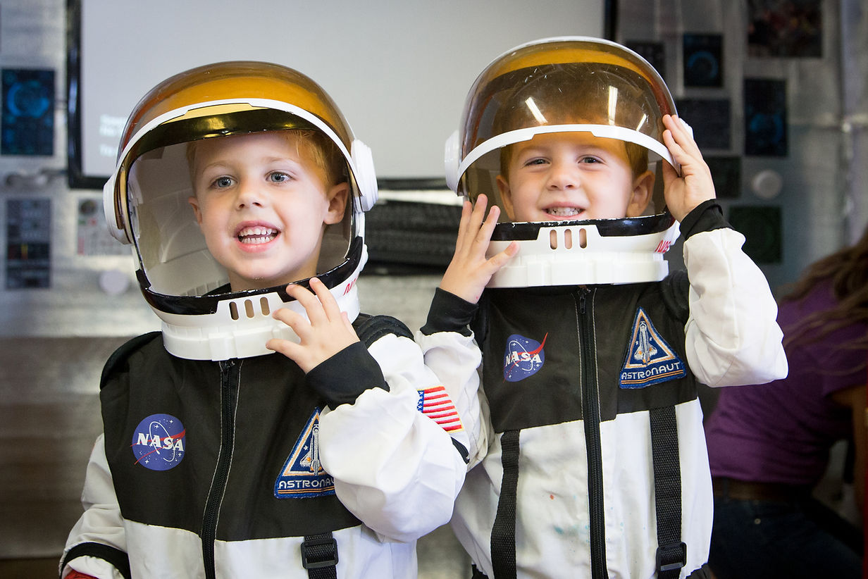Two children in astronaut suits and helmets smiling indoors, with a space-themed background.