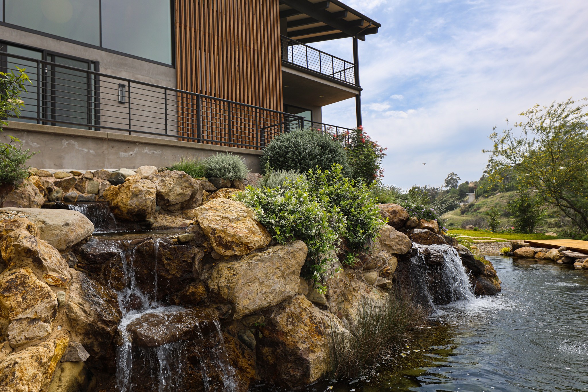 Water flows over rocks into a pond with trees and a building in the background.