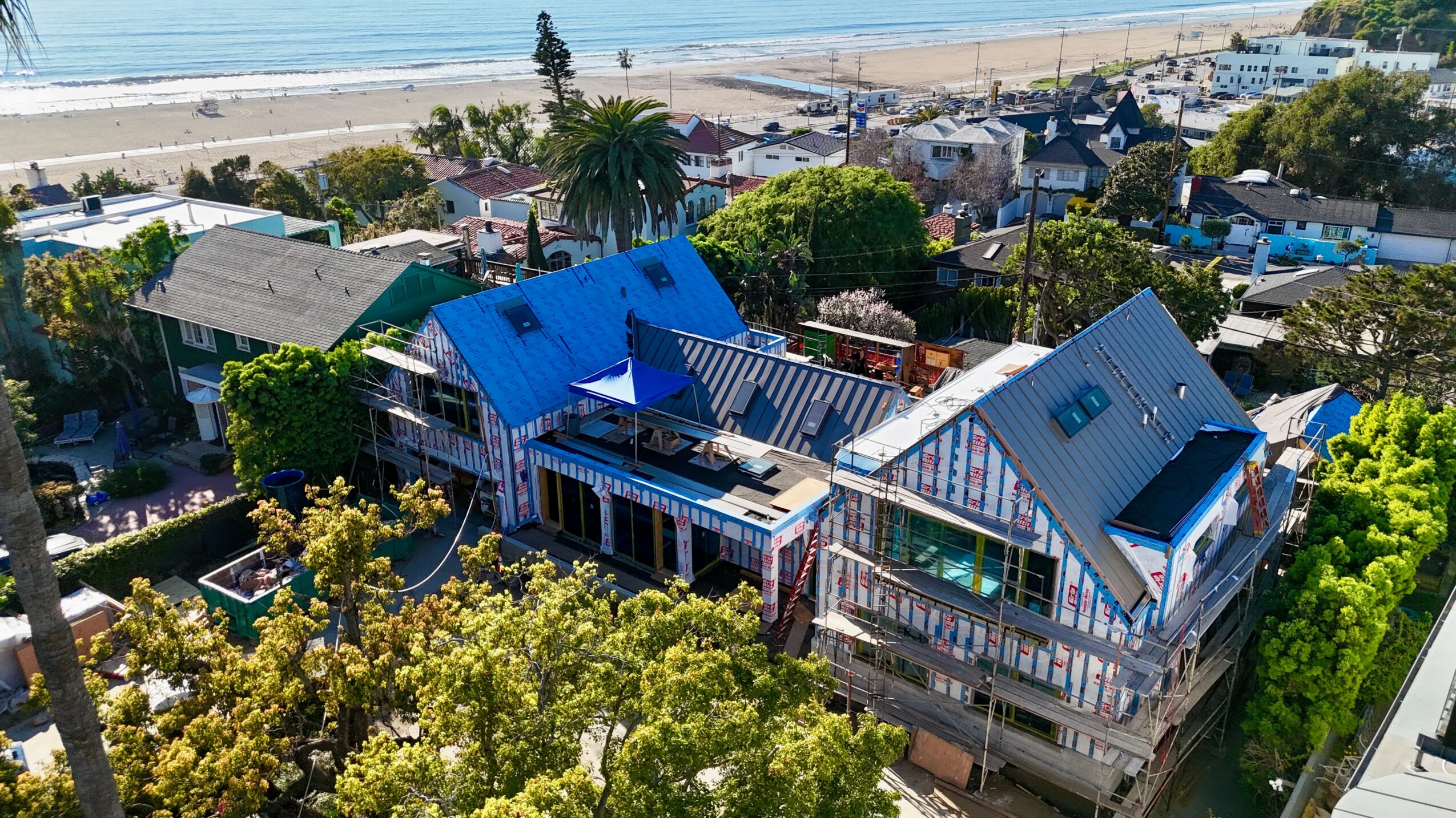 Aerial view of a coastal neighborhood with houses, trees, and a beach in the background.