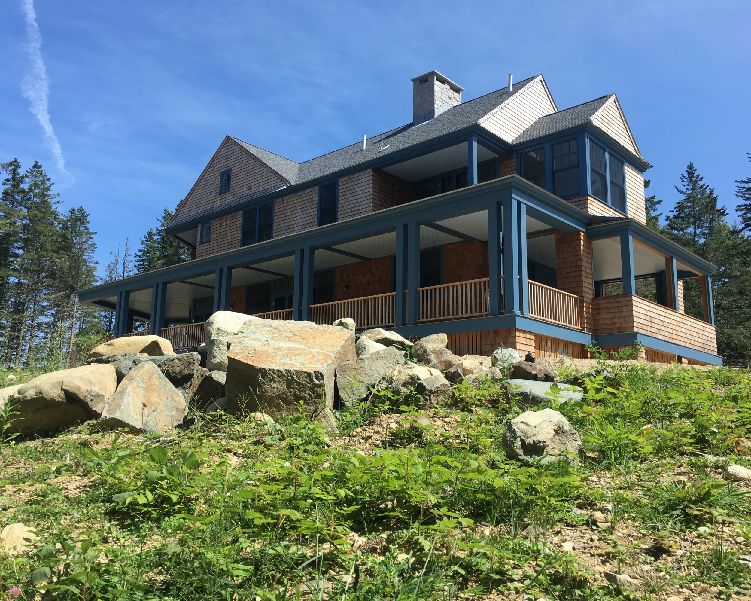 Large house with multiple levels, blue trim, surrounded by rocks and greenery, under a blue sky.