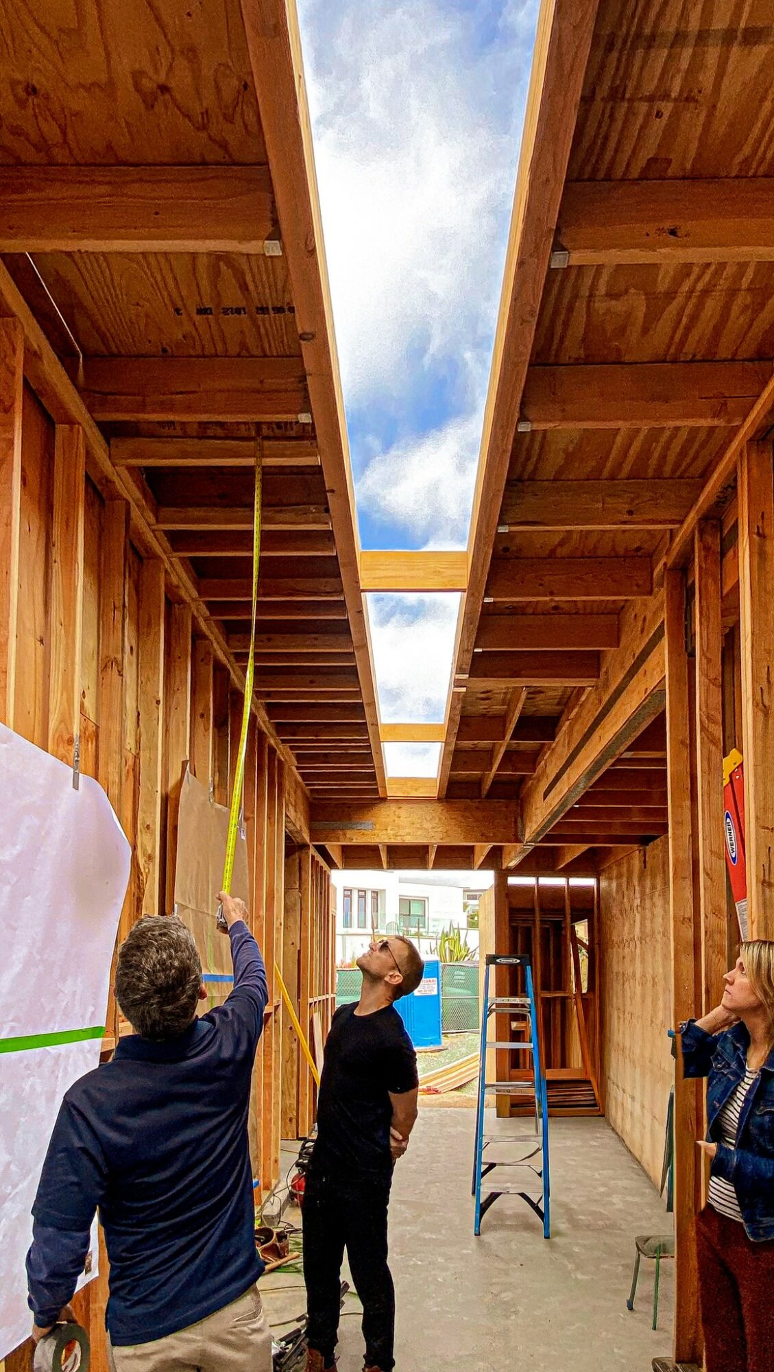 Four people inside a wooden structure, measuring ceiling height with a yellow tape measure, ladder nearby, open sky visible through ceiling opening.