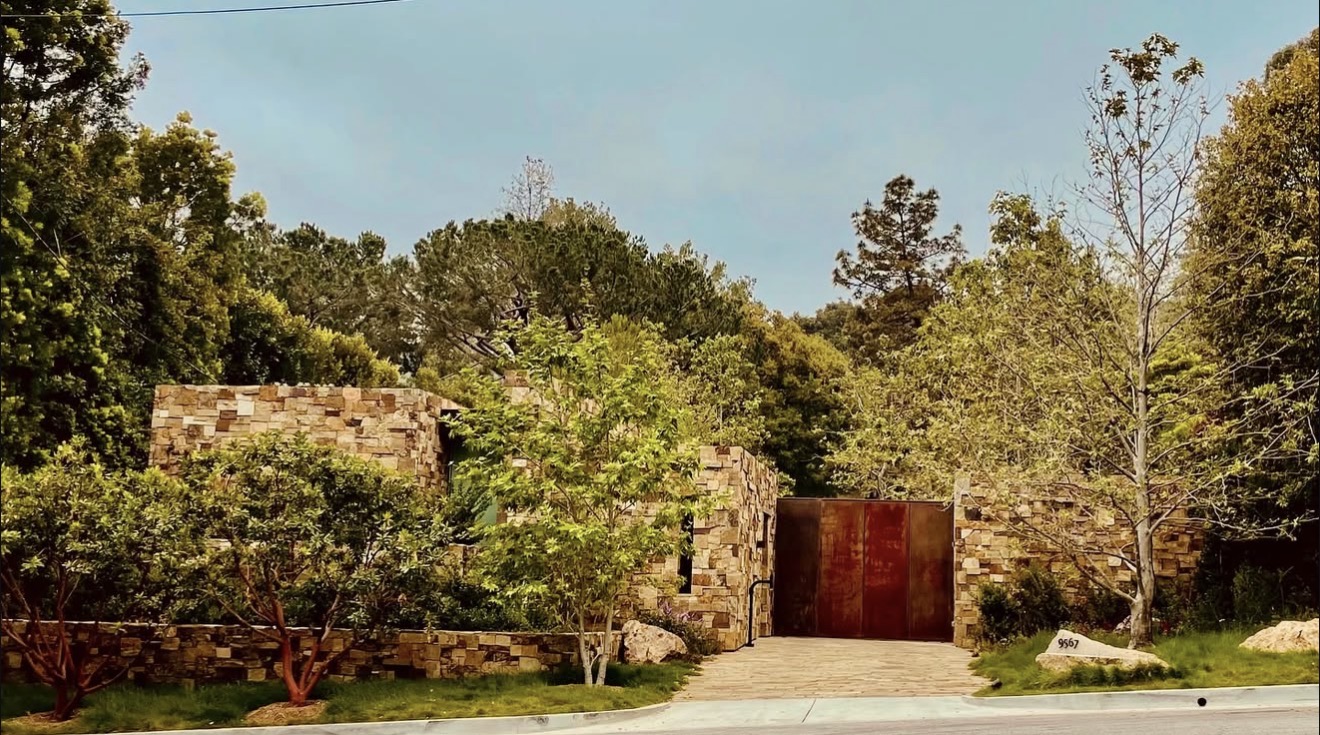 Stone wall with trees and greenery, driveway leading to a gate, clear sky in the background.