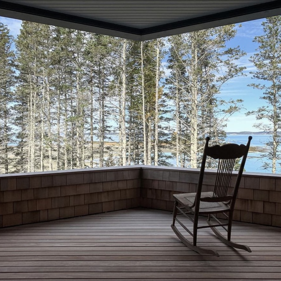 Rocking chair on porch overlooking trees and water, with blue sky and clouds.