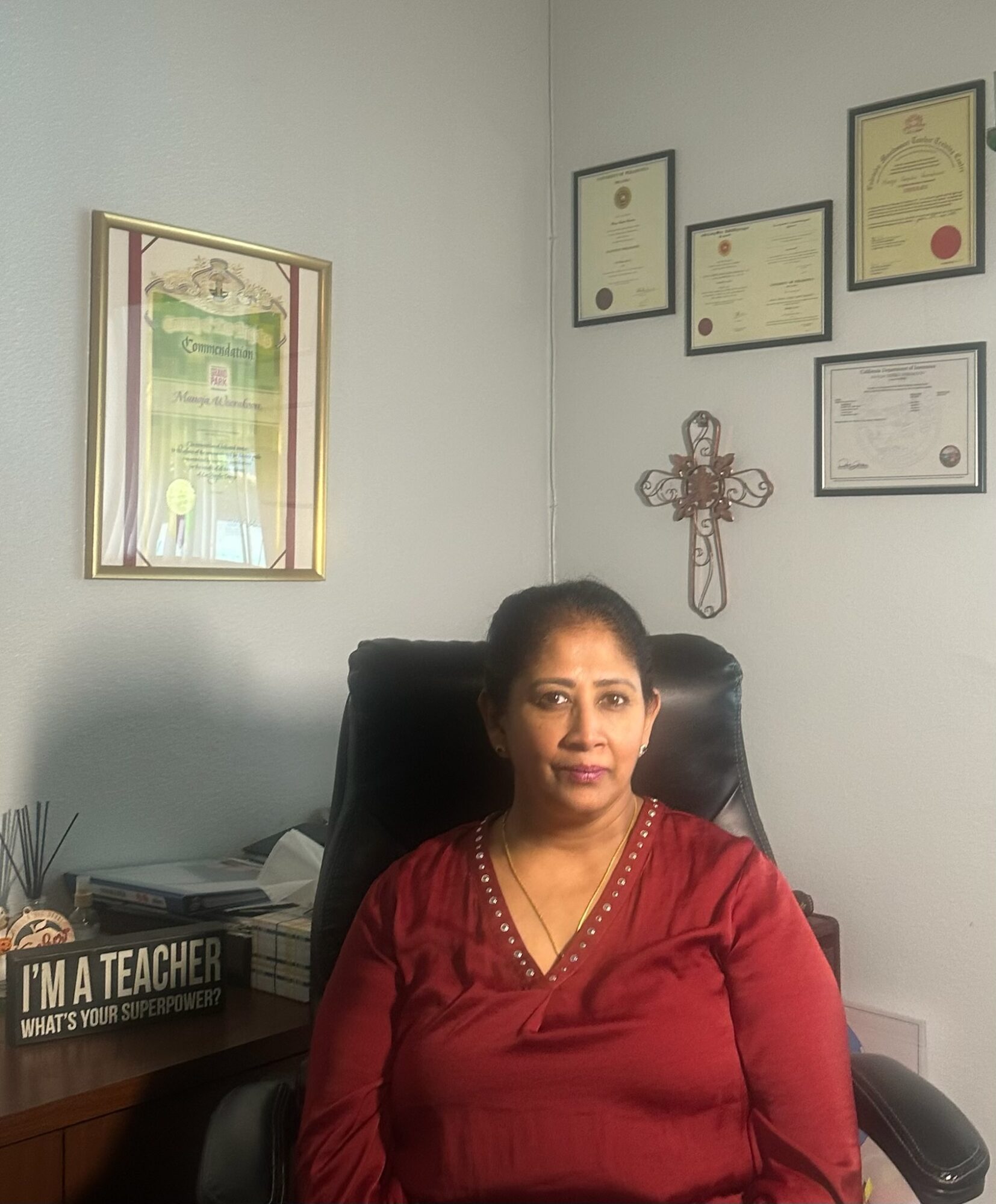 Woman sitting in a black chair in an office with framed certificates and a decorative cross on the wall.