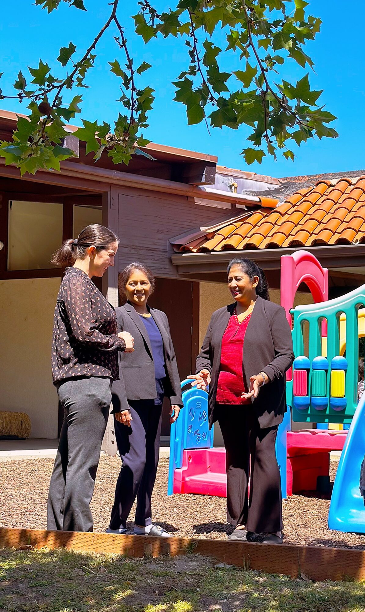 Four women stand outdoors near a colorful playground structure, talking and smiling, with a house and tree branches overhead.
