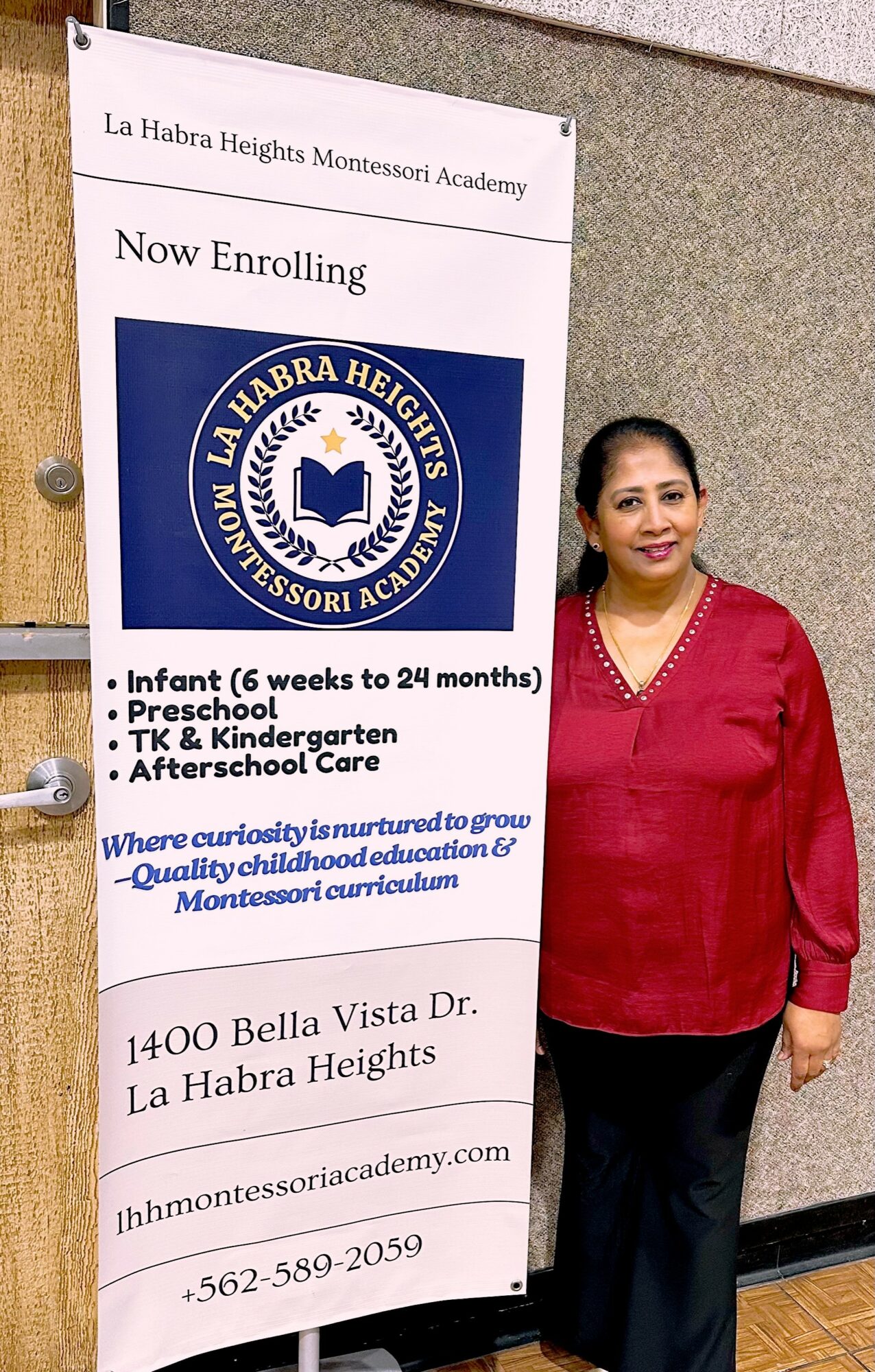 Woman in red top standing next to a large vertical sign for La Habra Heights Montessori Academy in a hallway.