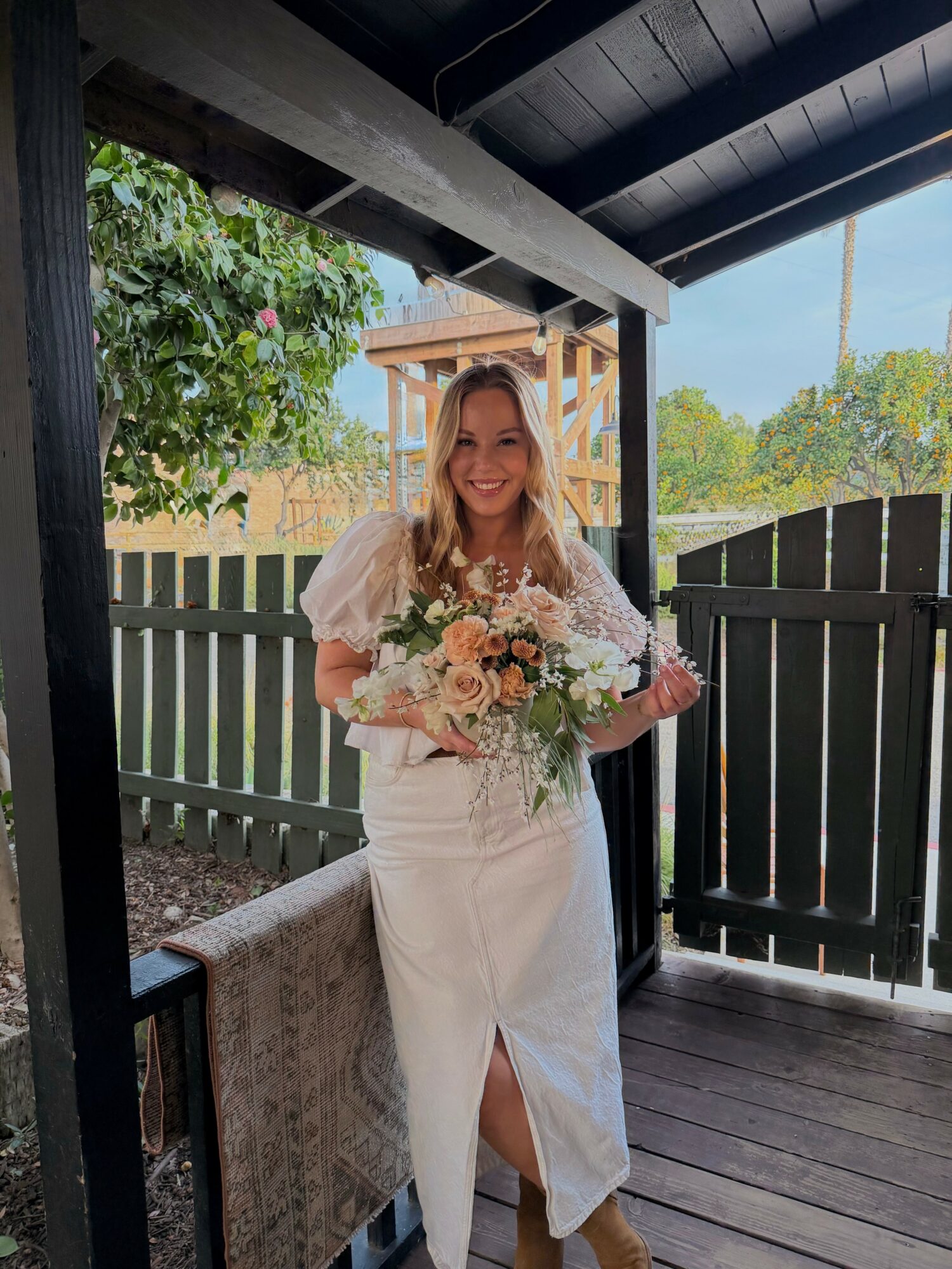 Woman in white dress holding a bouquet, standing on a porch with a wooden fence and garden background.