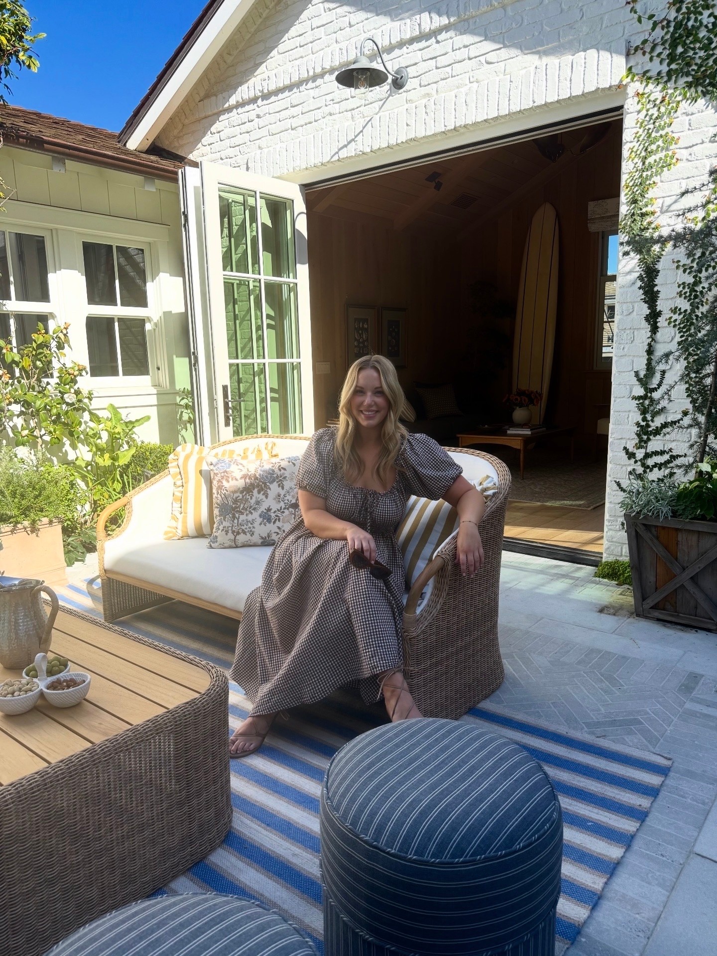 Woman sitting on outdoor sofa with patterned dress, smiling, in front of open garage door, with house and plants nearby.
