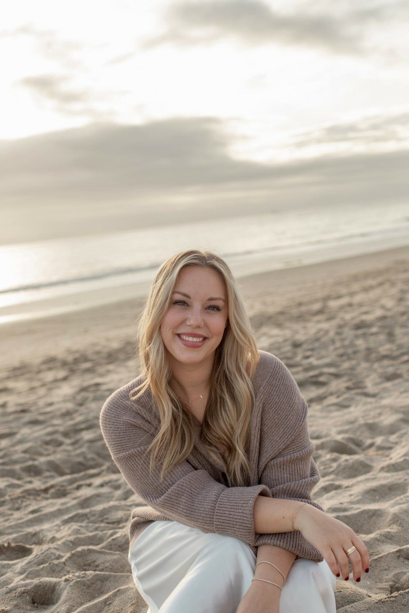 Woman with long blonde hair smiling on a beach during sunset, wearing a brown sweater and white pants.