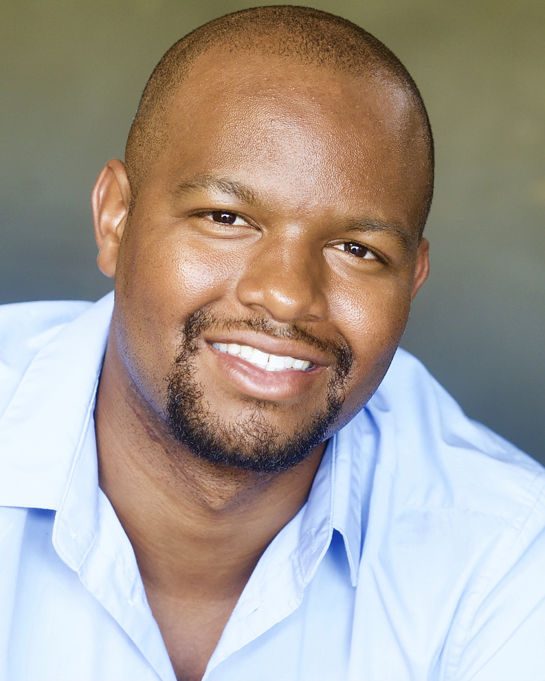 Close-up portrait of a smiling man with a shaved head and beard, wearing a light blue shirt.