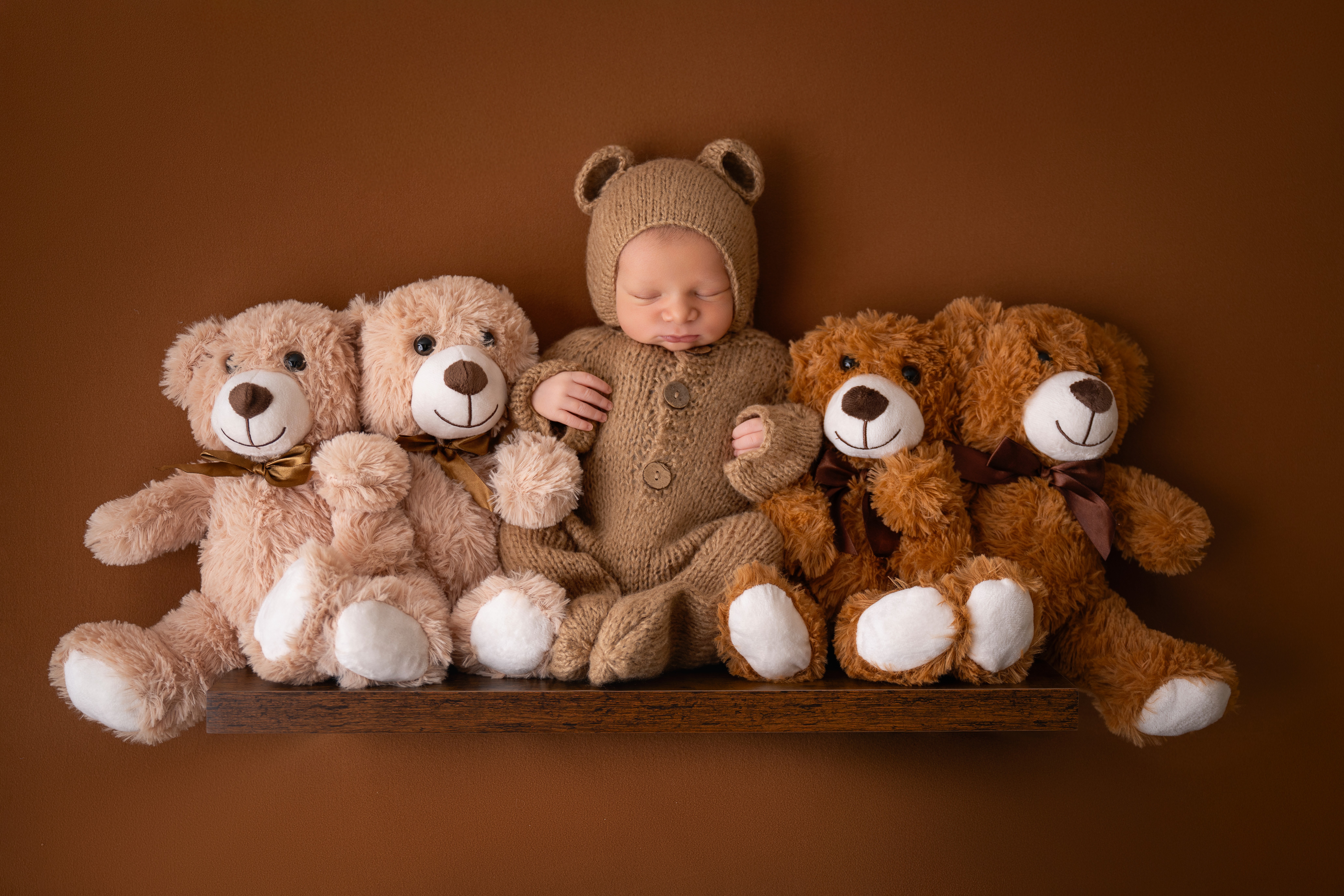 Baby dressed in bear costume with four teddy bears on a shelf against a brown background.