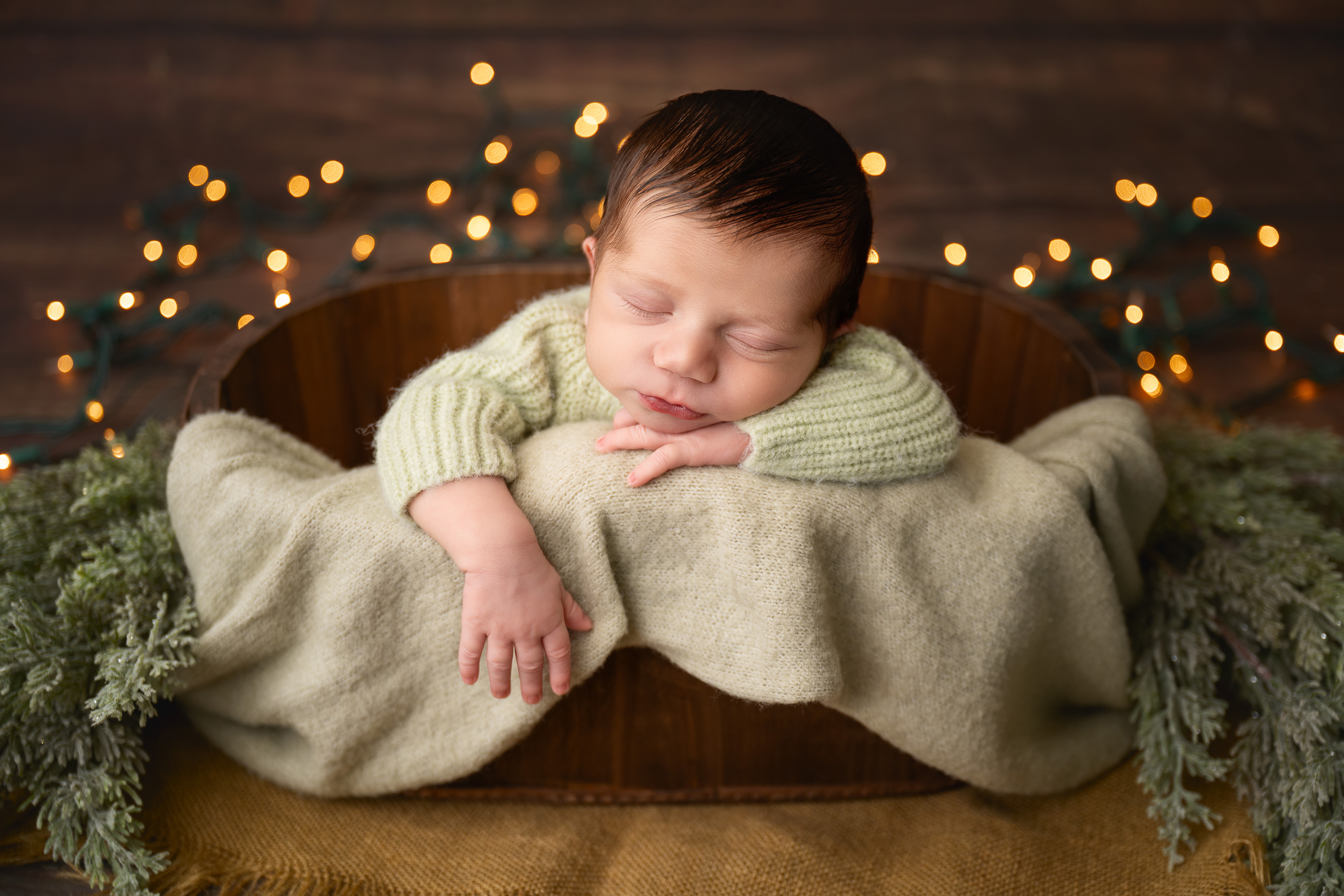 Baby sleeping with head resting on folded arms, lying on a blanket in a cozy setting with blurred lights in background.