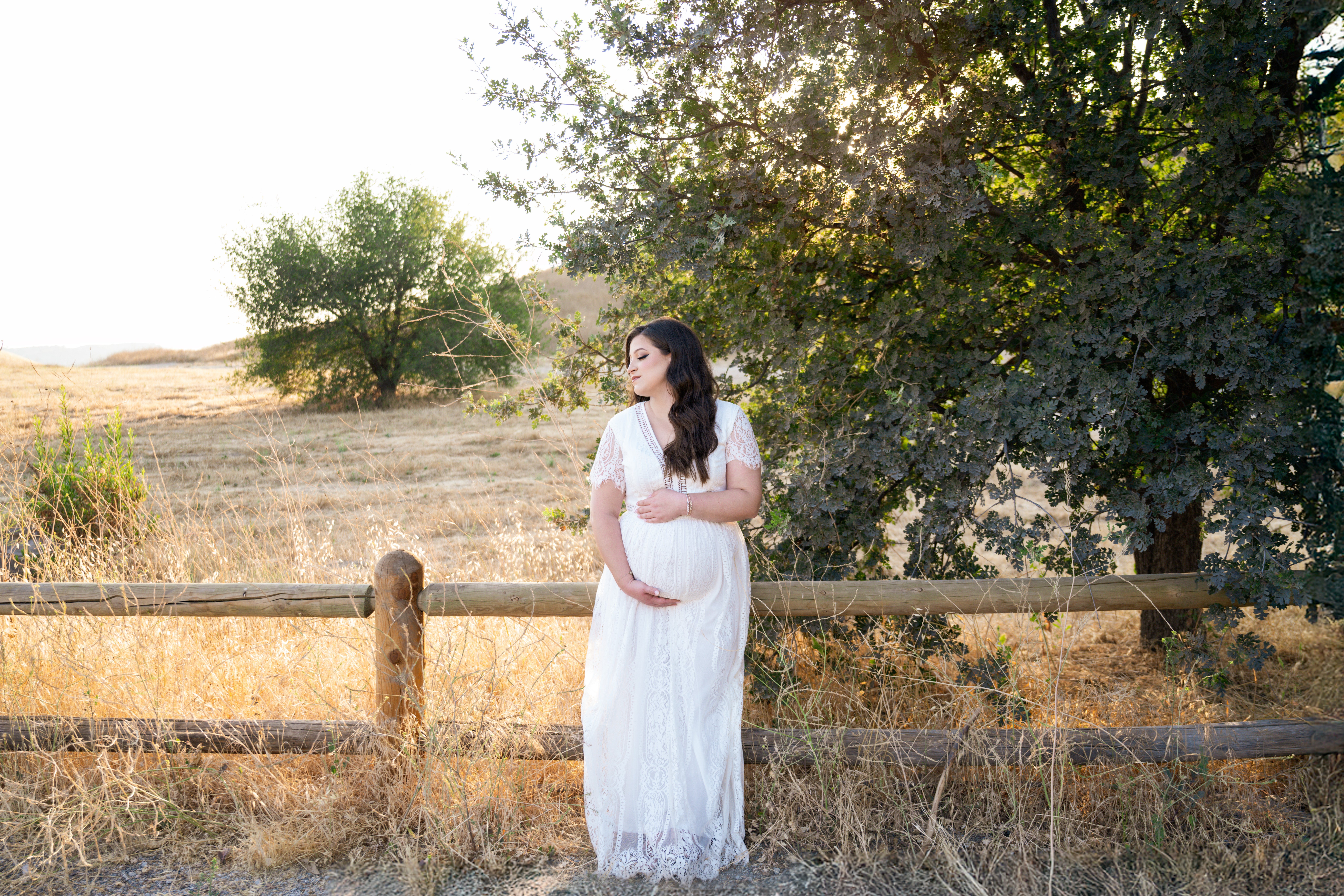Pregnant woman in a white dress standing near a wooden fence in a field with trees and sunlight.