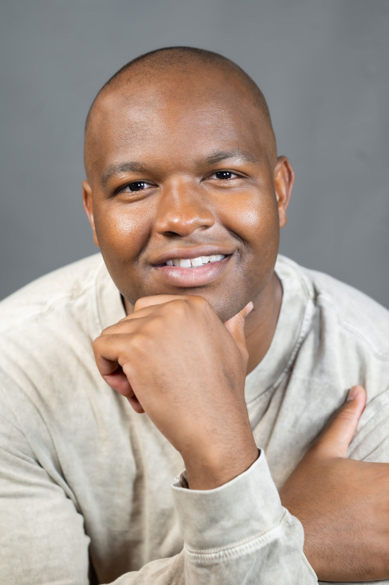 Man with a shaved head smiling, resting chin on hand, wearing a light-colored shirt, gray background.