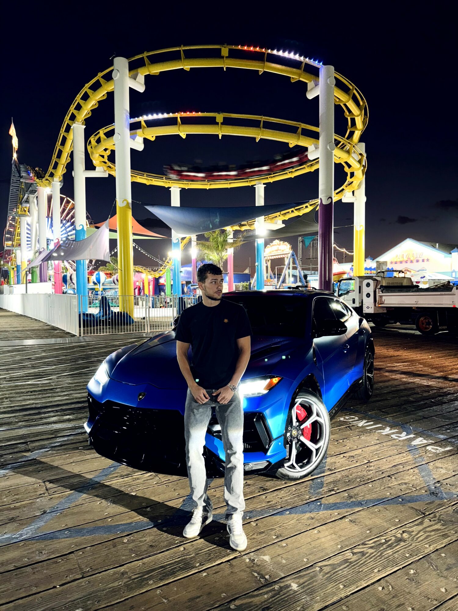 Person standing in front of a blue sports car at night with a colorful amusement park ride in the background.