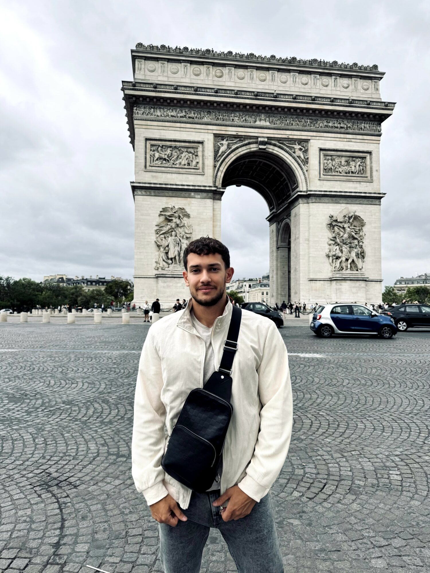 Young man standing in front of Arc de Triomphe, wearing a white jacket and carrying a black bag.
