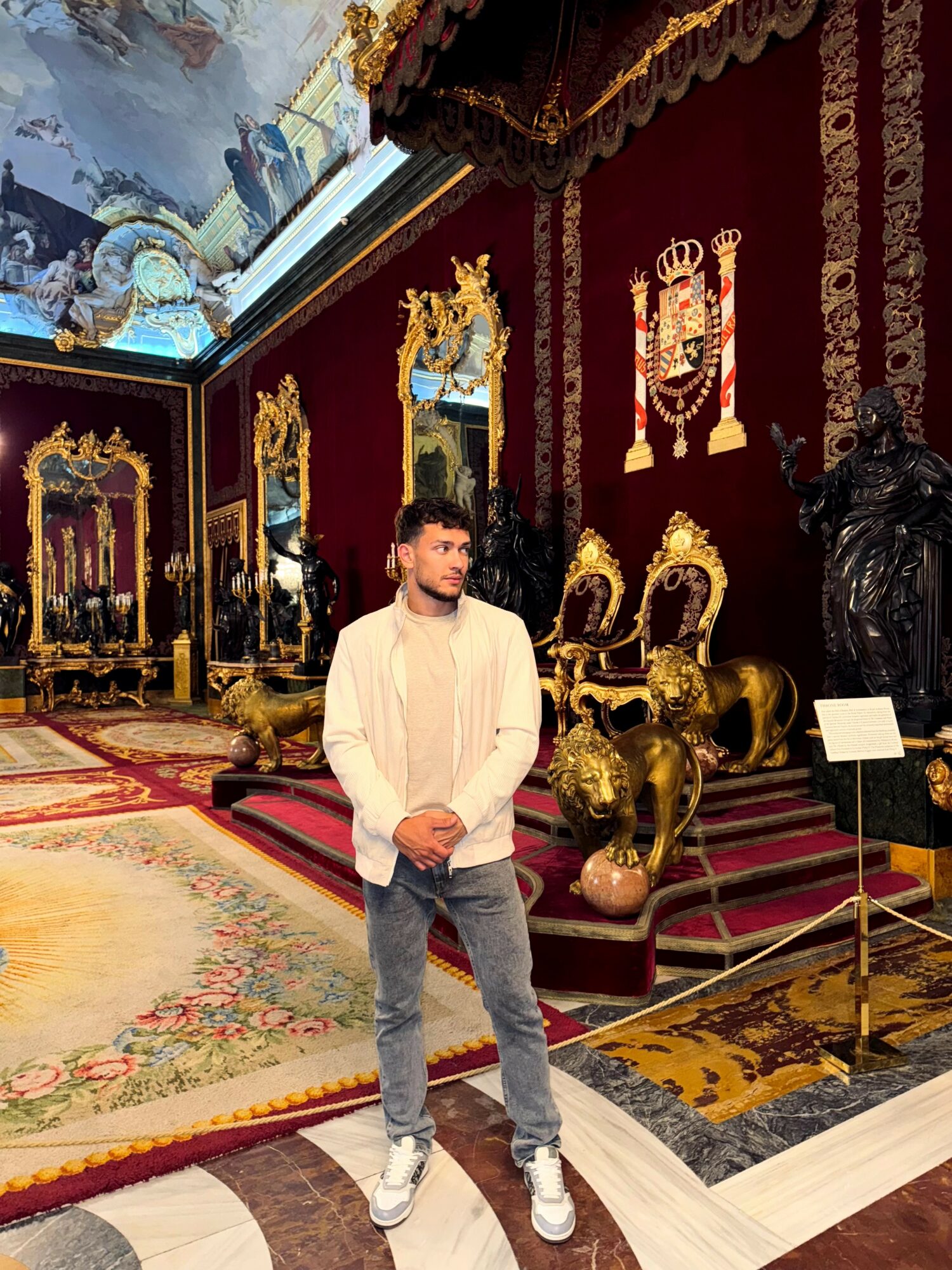 Young man in casual clothing standing in ornate room with gold furniture and decorative wall art.