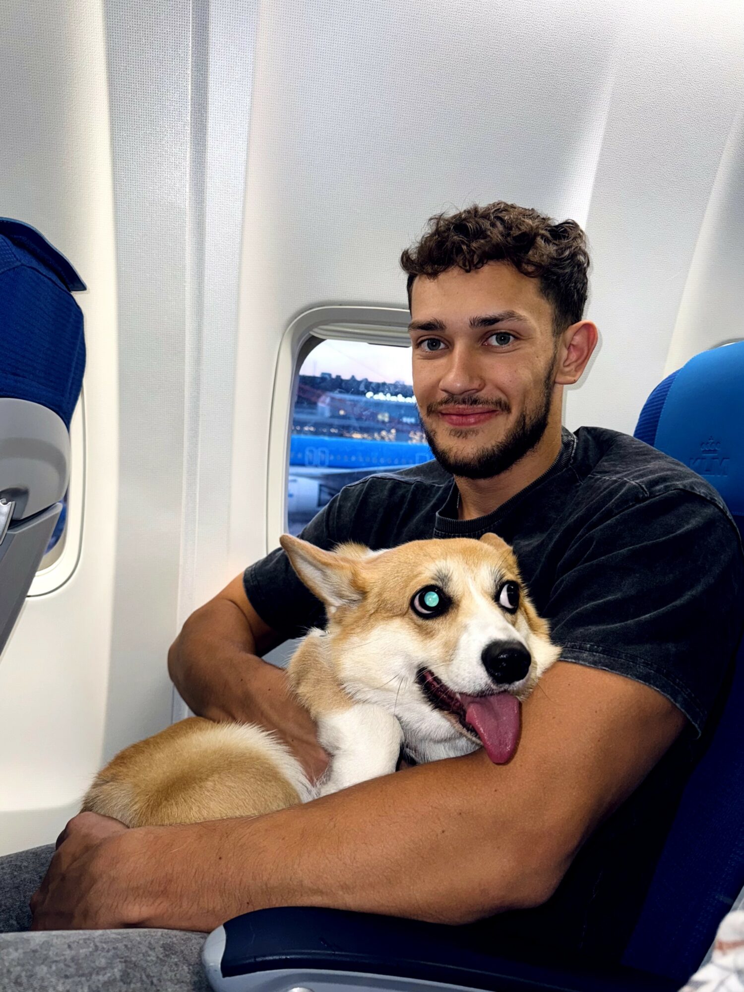 Man holding a dog with blue eyes on an airplane seat, window view outside, smiling.