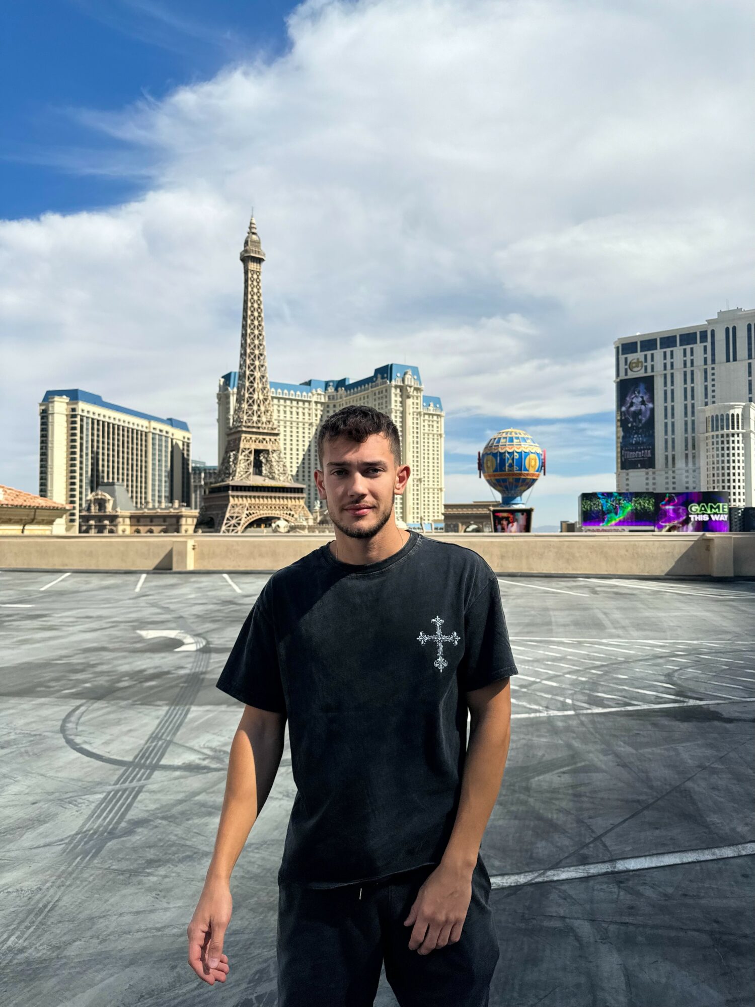 Young man standing outdoors with cityscape, including the Eiffel Tower replica and a globe sculpture, in the background.