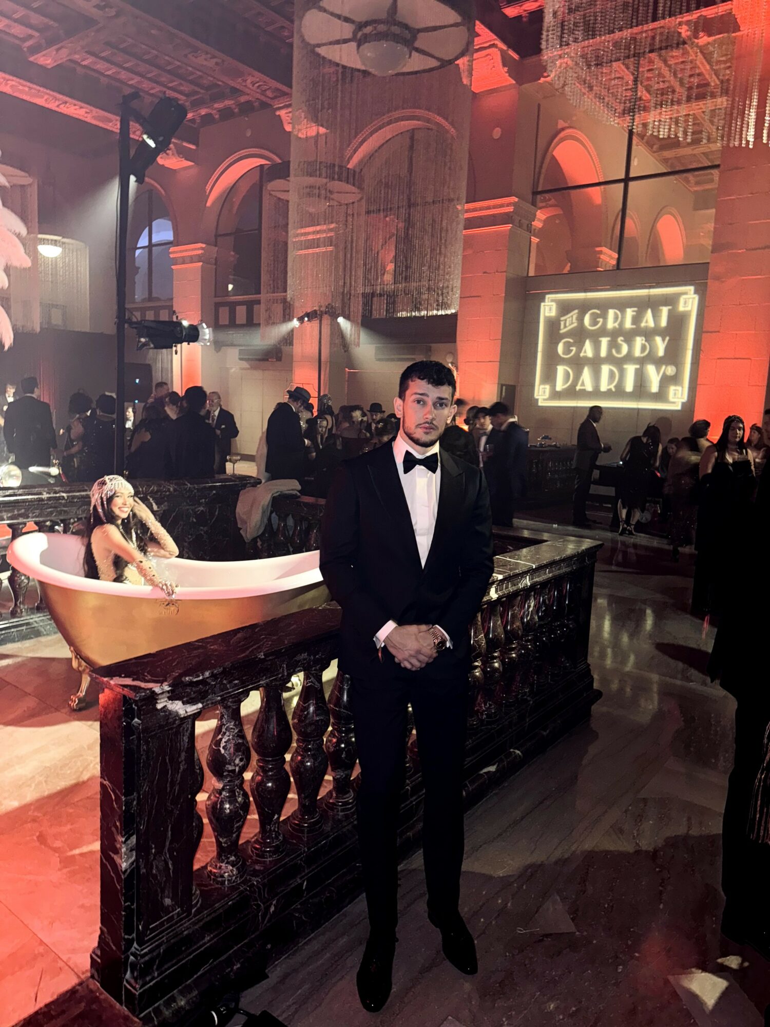 Man in tuxedo standing in a decorated event space with a bathtub and a large illuminated sign in the background.