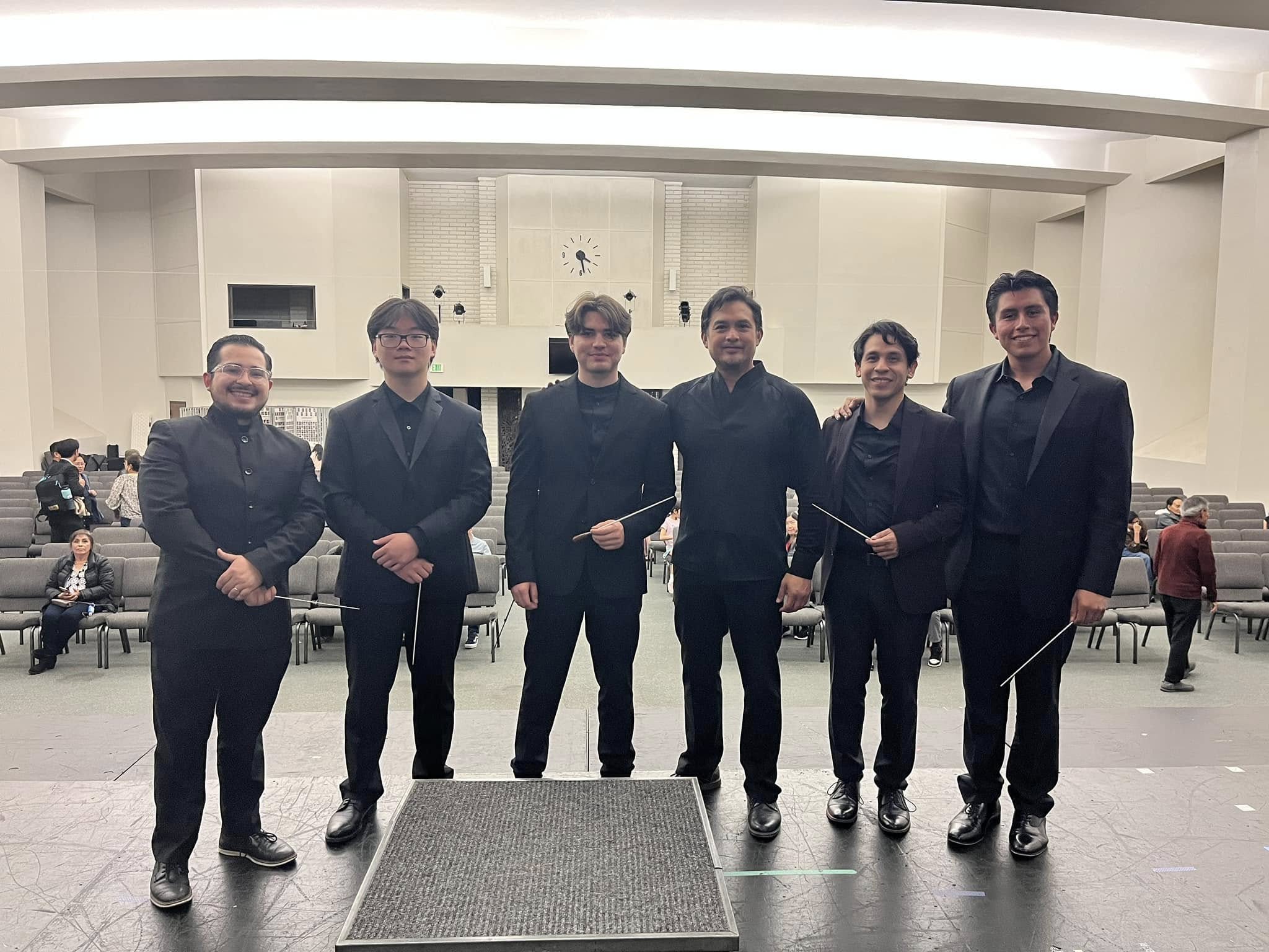 Six people in formal attire holding batons, standing in a large indoor hall with a stage and audience in background.