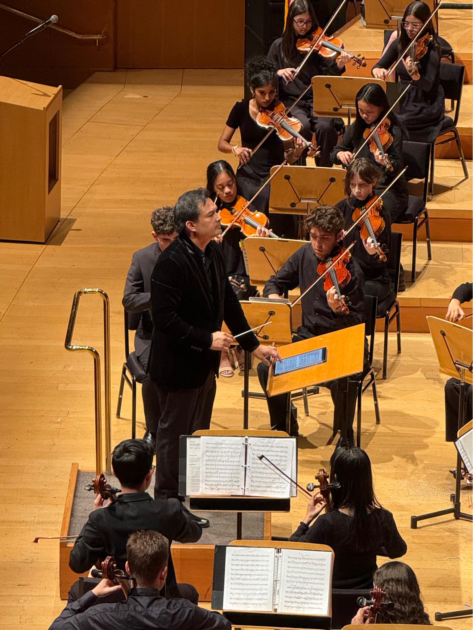 Orchestra conductor leading musicians on stage with string instruments, music stands, and sheet music, in a concert hall.
