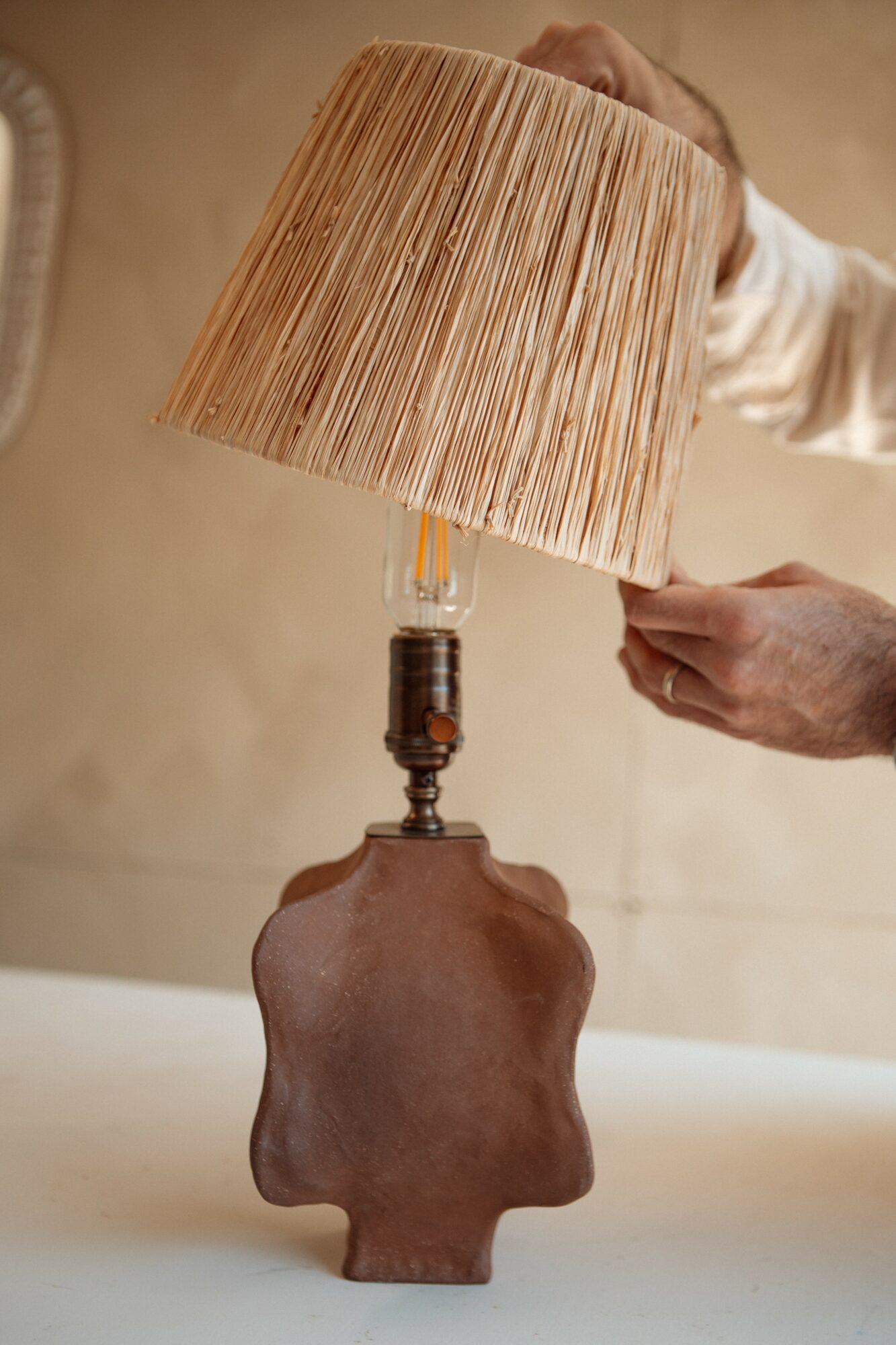 Person adjusting a lamp with a beige lampshade and a brown ceramic base.