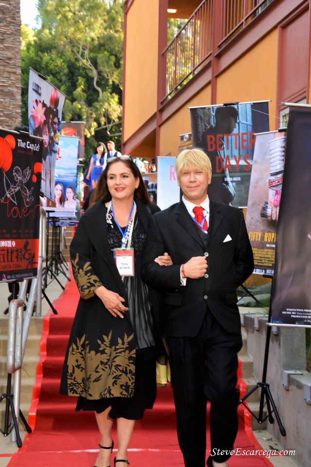 Two people, a woman and a man, walking on a red carpet outdoors, with posters and a building in the background.