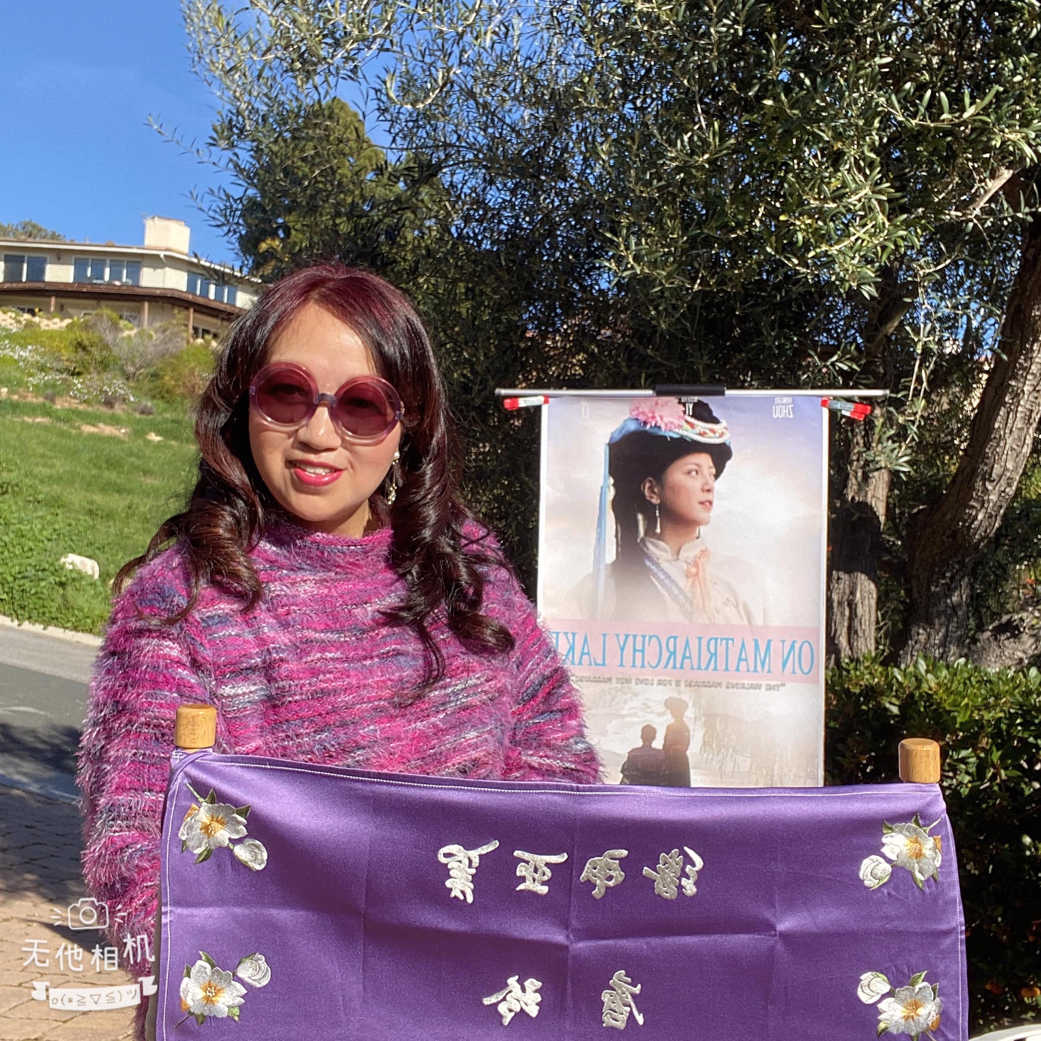 Woman with sunglasses and wavy hair standing outdoors, holding a large purple banner with Chinese characters and flowers, behind a poster of a woman.