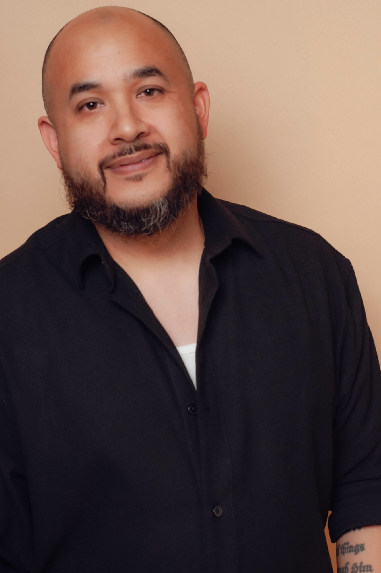 Man with a beard and shaved head wearing a black shirt, smiling, against a beige background.