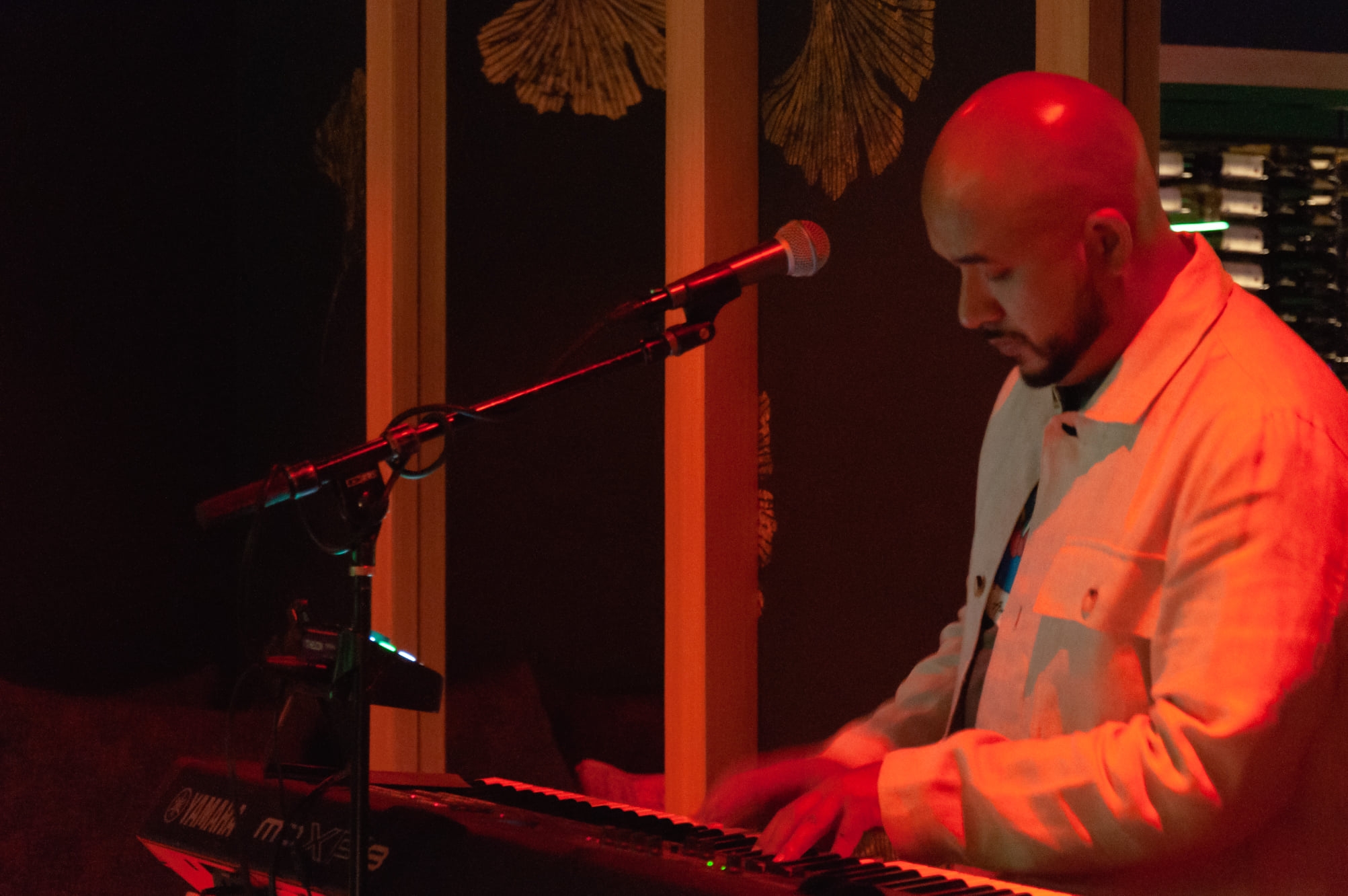 Bald man playing keyboard on stage with microphone, illuminated by red lighting, in a dark setting.