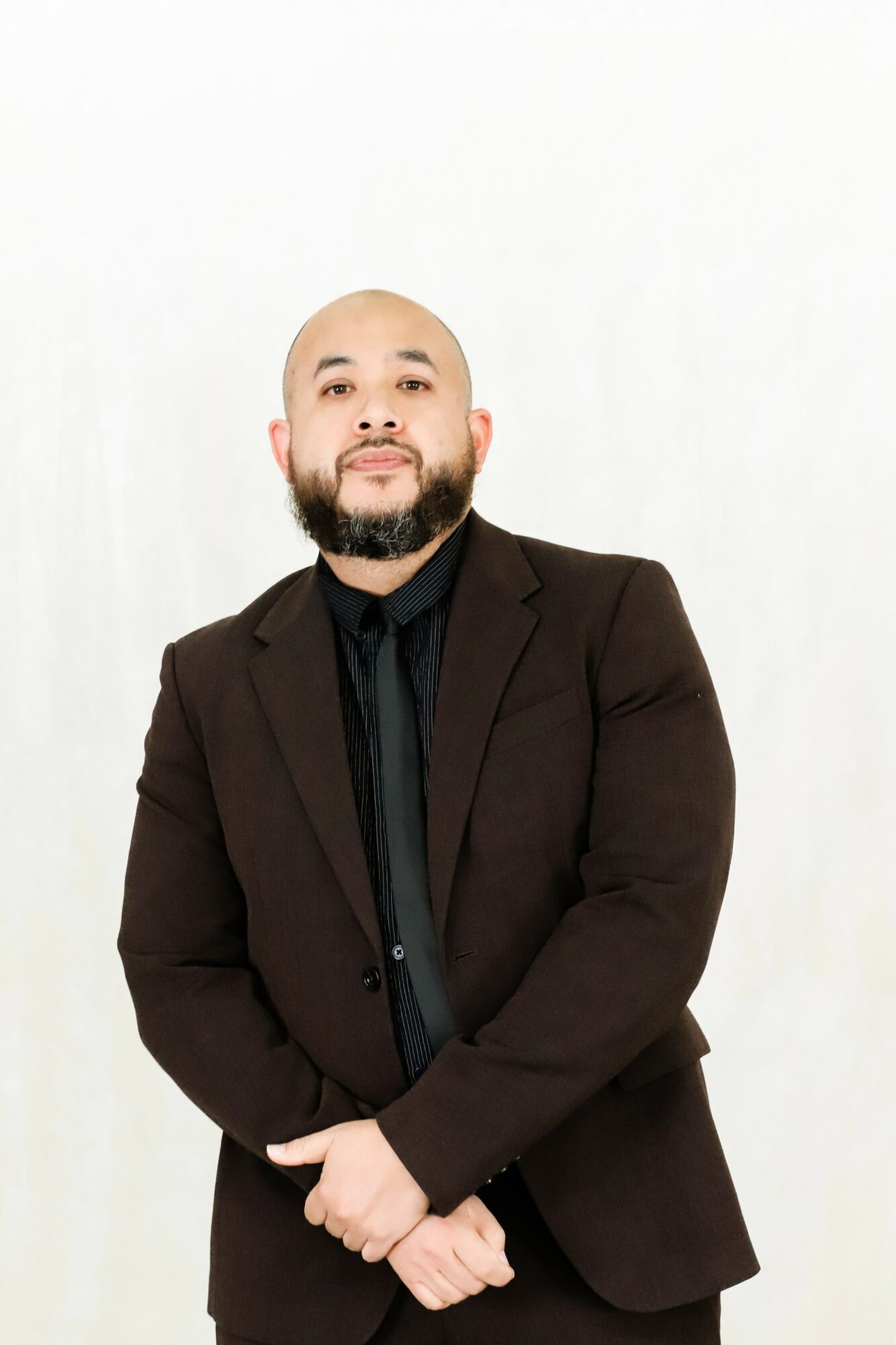Man with a beard in a black suit and shirt, standing against a white background.