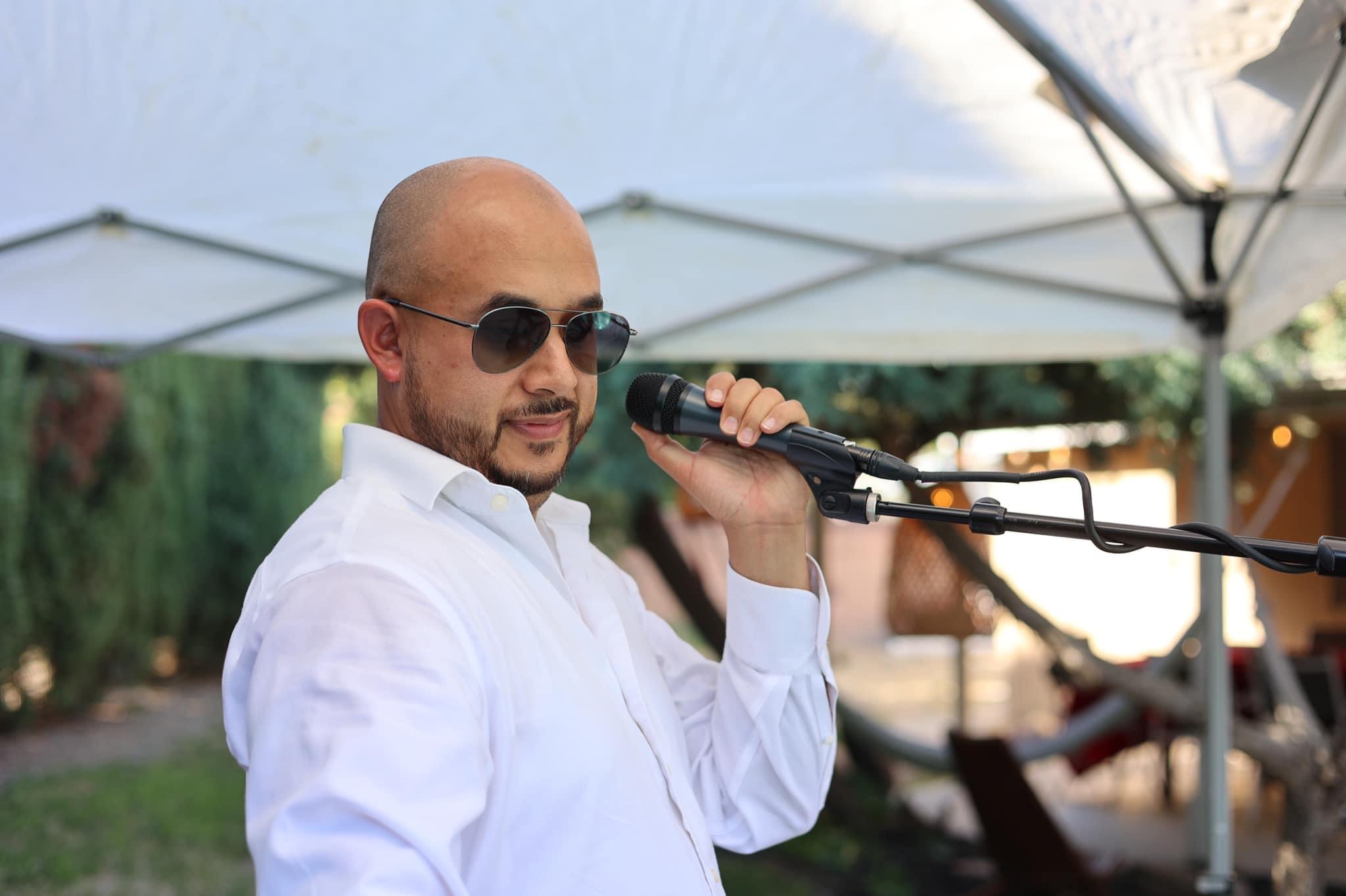 Man with sunglasses and a white shirt holding a microphone outdoors under a canopy.