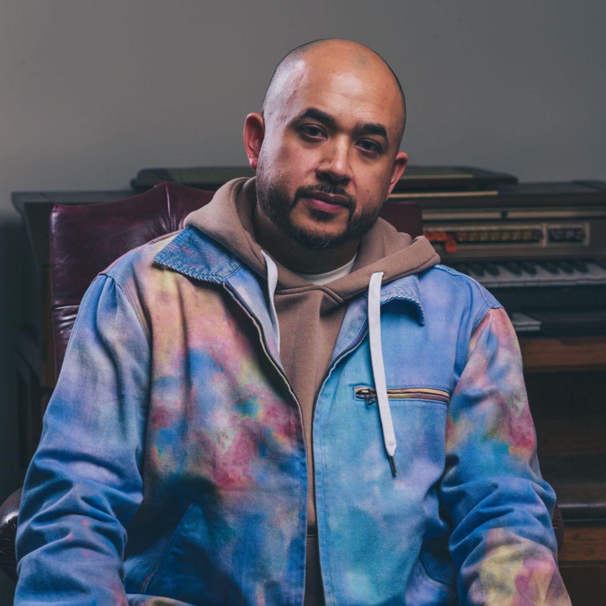 Man with a beard and shaved head sitting in front of a keyboard, wearing a colorful jacket and hoodie.
