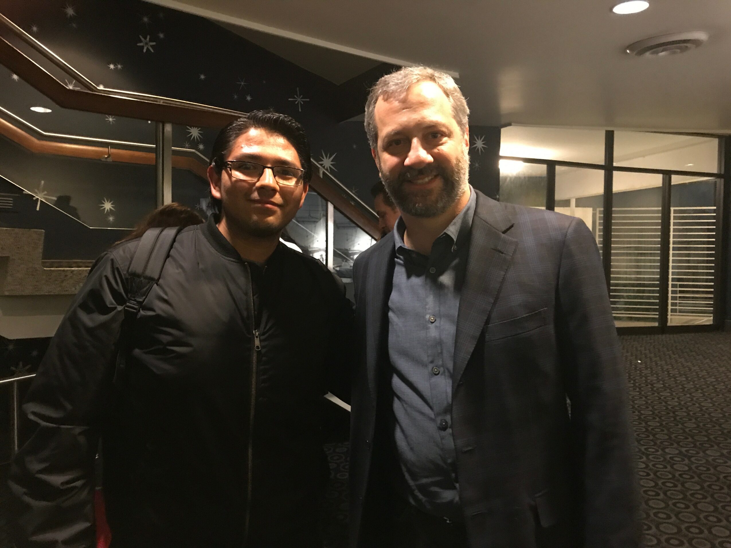 Two men standing indoors, one wearing glasses and a black jacket, the other in a blazer and shirt, smiling.