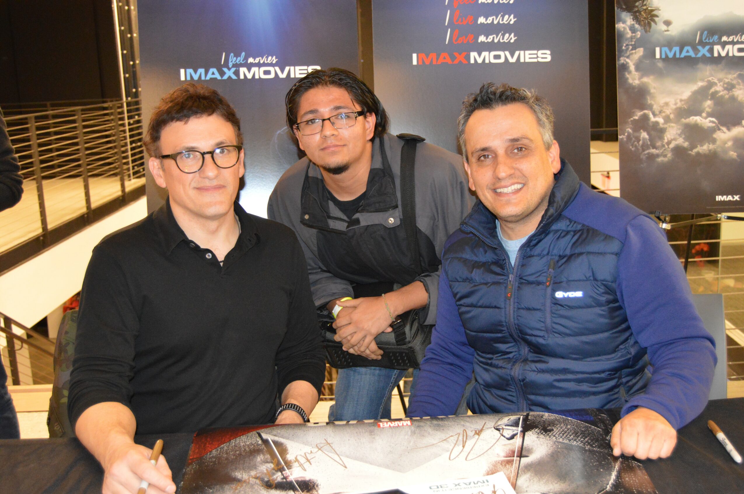 Three men sitting at a table with promotional posters in the background, smiling at the camera.