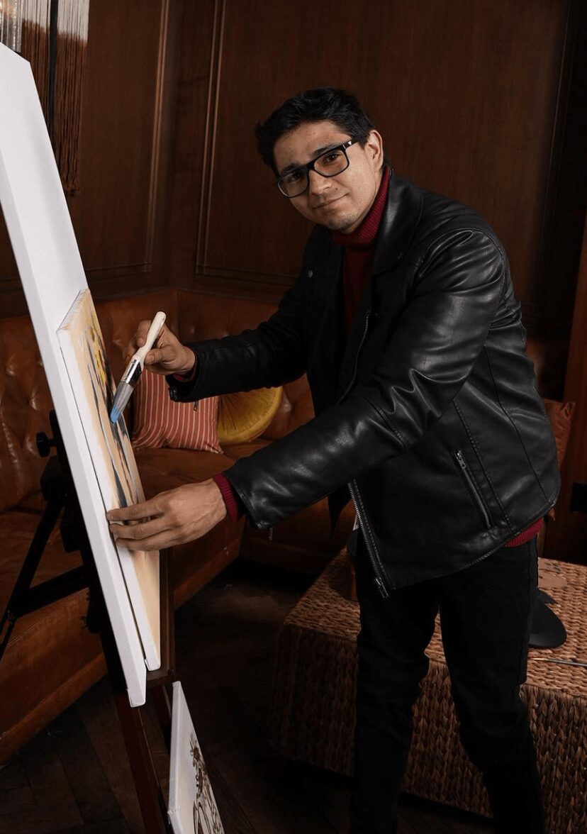 Person with glasses and dark hair, wearing a black leather jacket, painting on a canvas in a room with wooden walls.