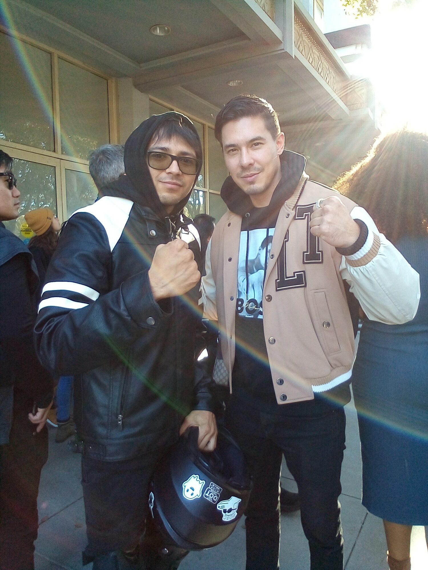 Two young men pose outdoors, one holding a helmet, both smiling and wearing jackets, with sunlight shining behind them.