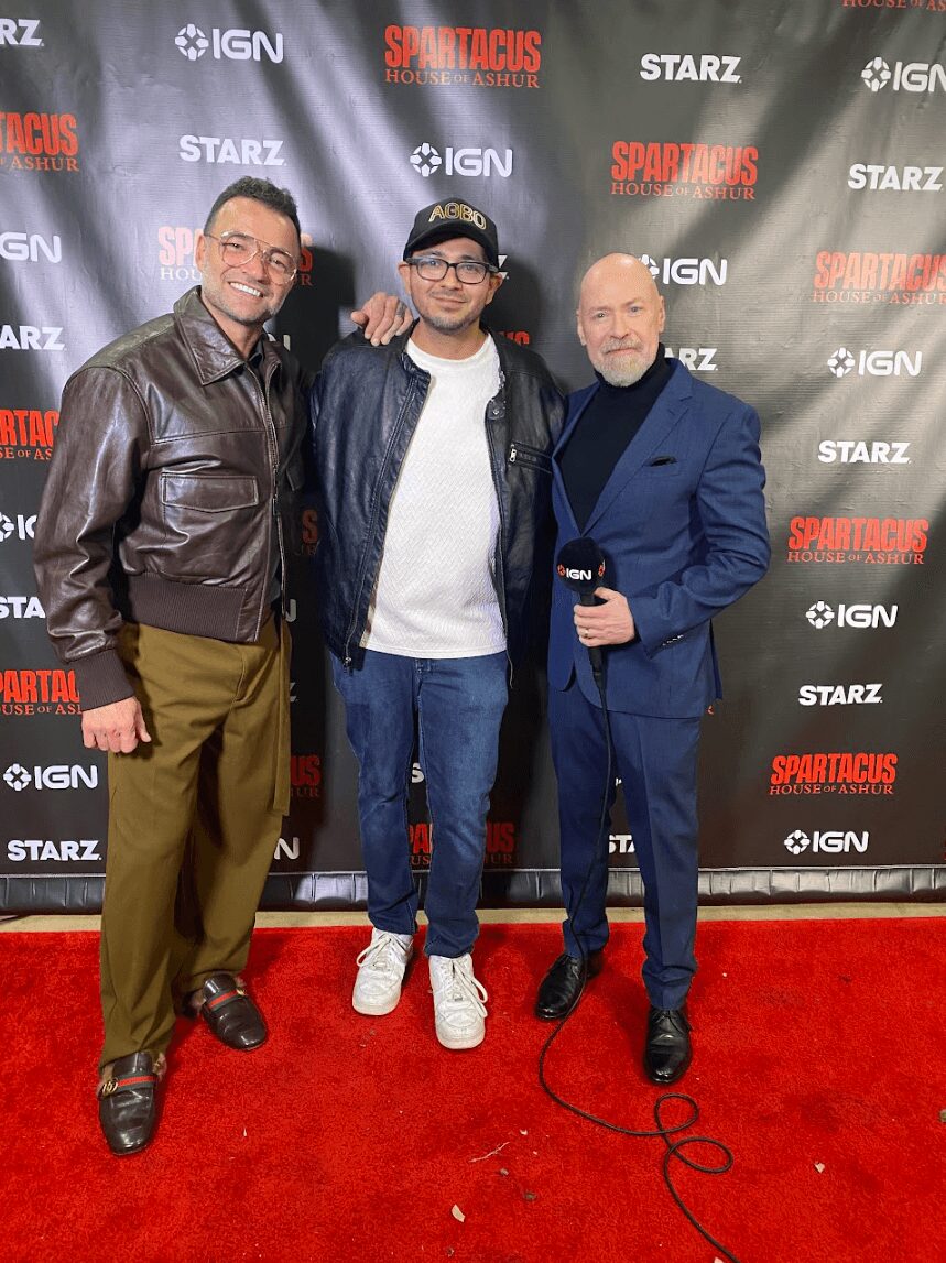 Three men standing on red carpet in front of a black backdrop with logos, smiling, one holding a microphone.