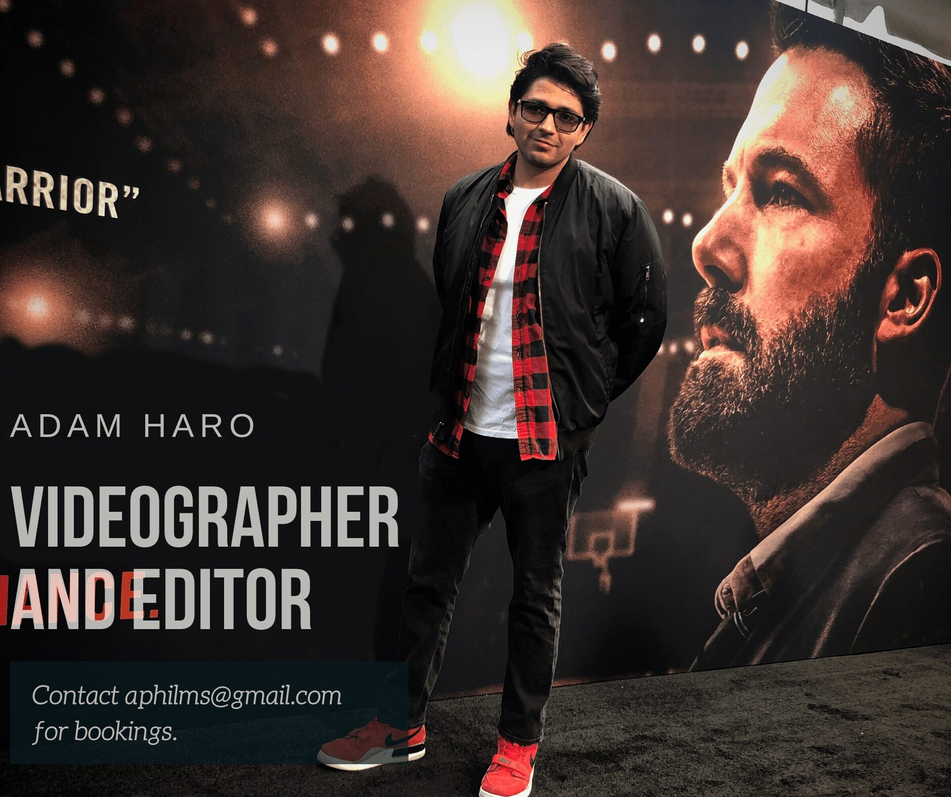 Young man standing in front of a large poster with a bearded man's face, wearing glasses, a black jacket, and red shoes.