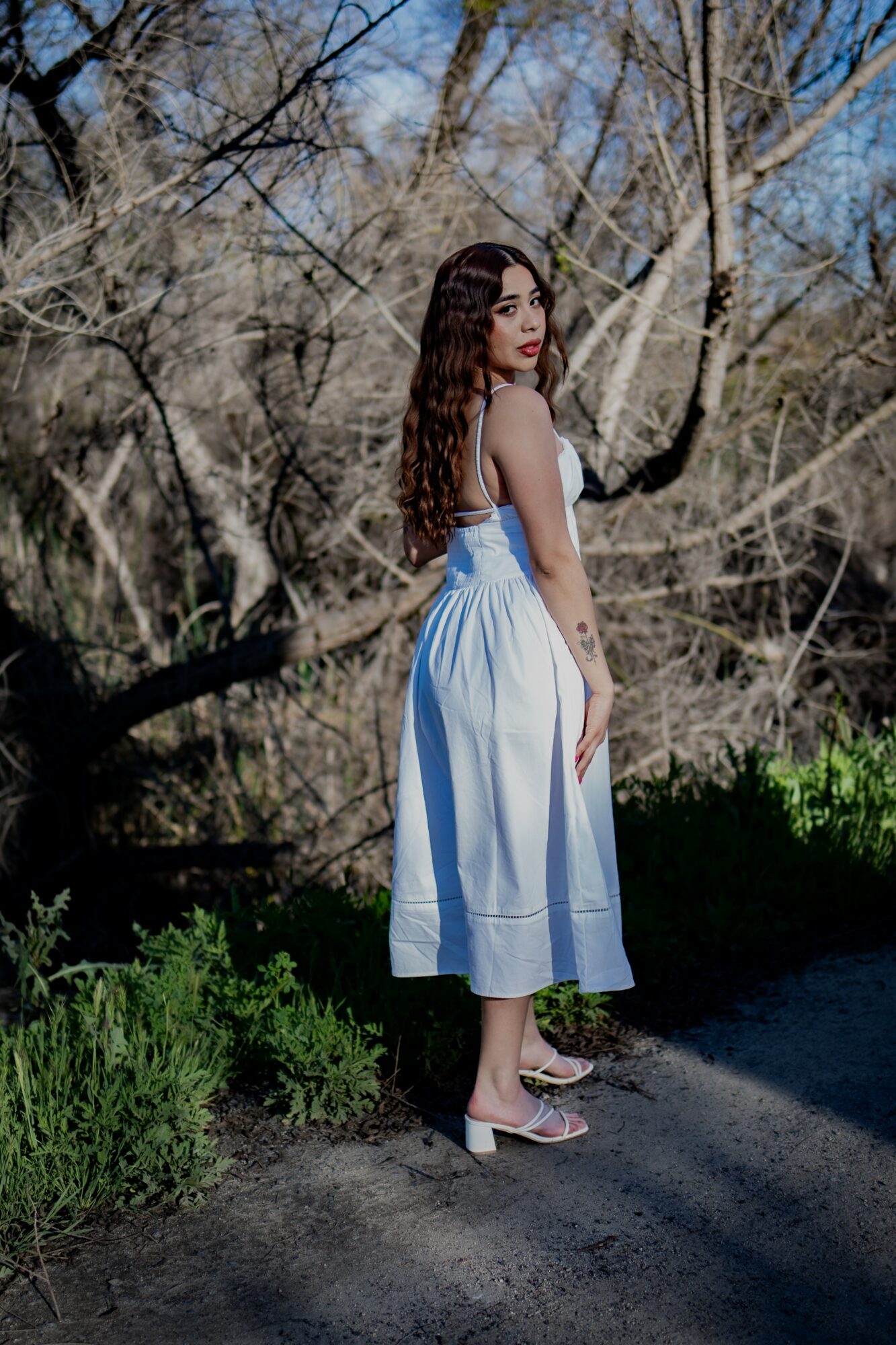 Young woman with long dark hair in a white dress standing outdoors among leafless trees and greenery.