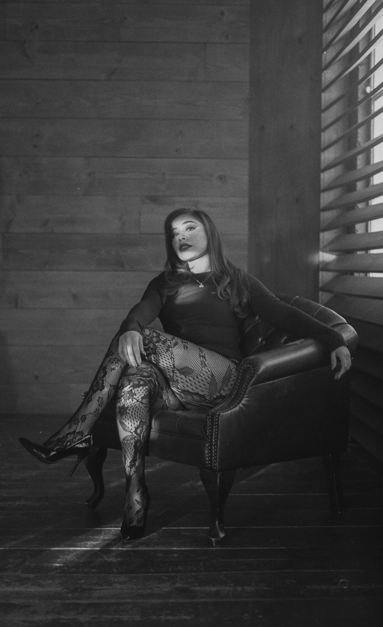 Woman sitting on a chair in a room with wooden walls and window blinds, wearing dark clothing and patterned tights.