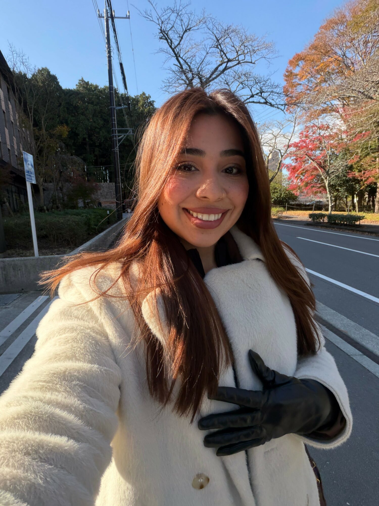 Young woman with long brown hair smiling outdoors in autumn, wearing a white coat and black gloves.