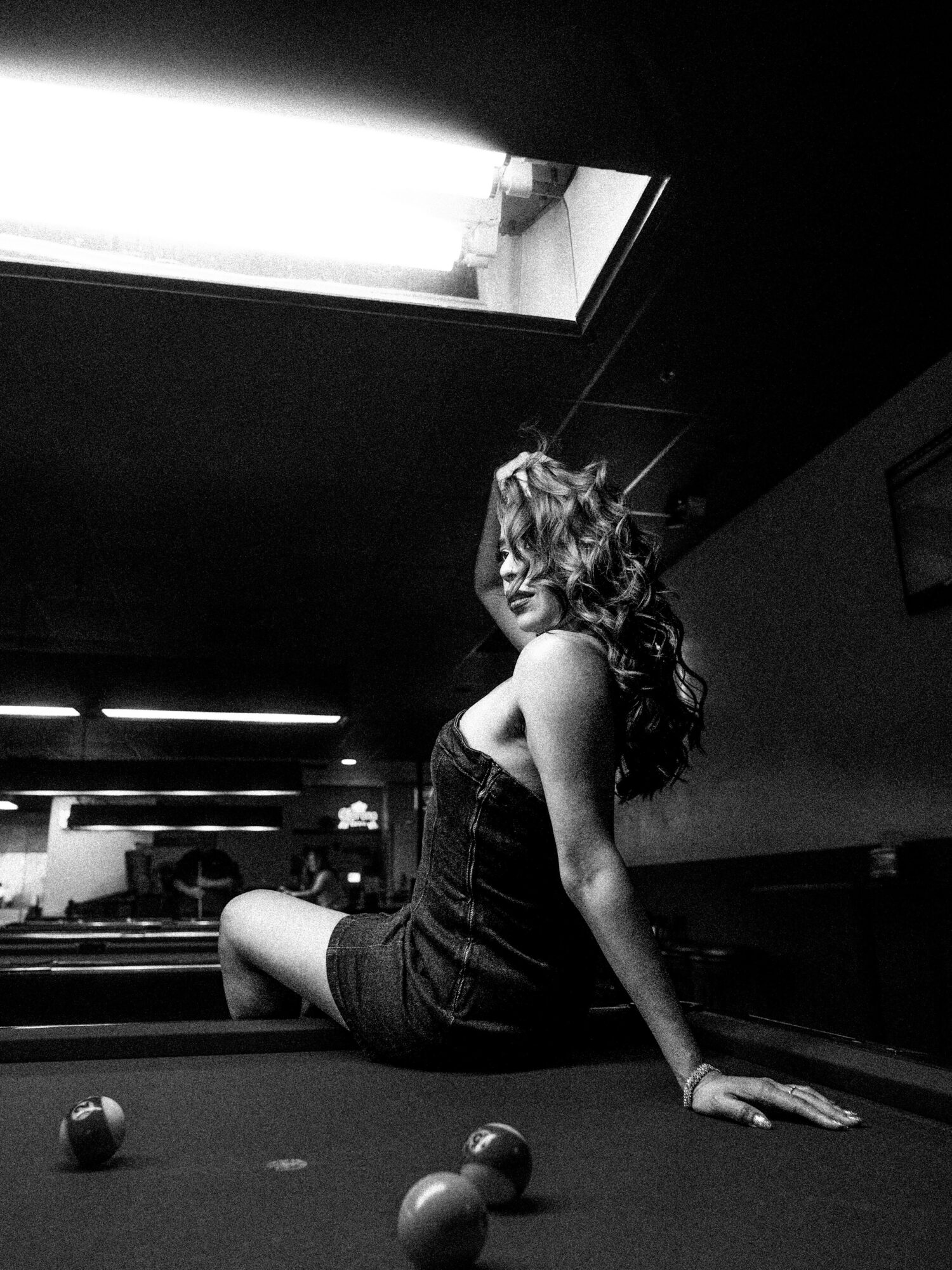 Woman with long curly hair sitting on a pool table, looking away, with pool balls nearby, in a dimly lit room.