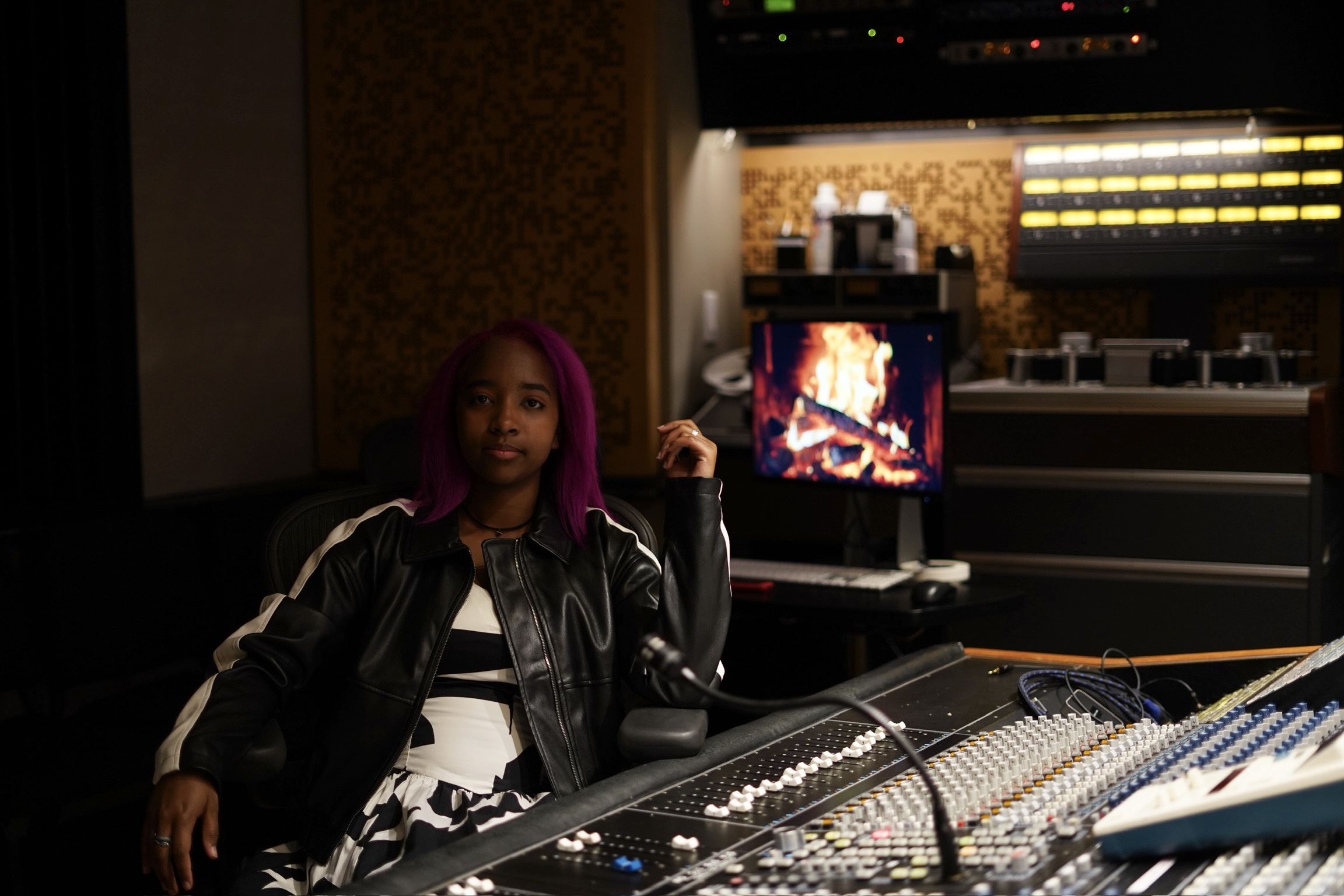 Woman with purple hair sitting in a recording studio with a mixing console and a computer displaying a fire image.
