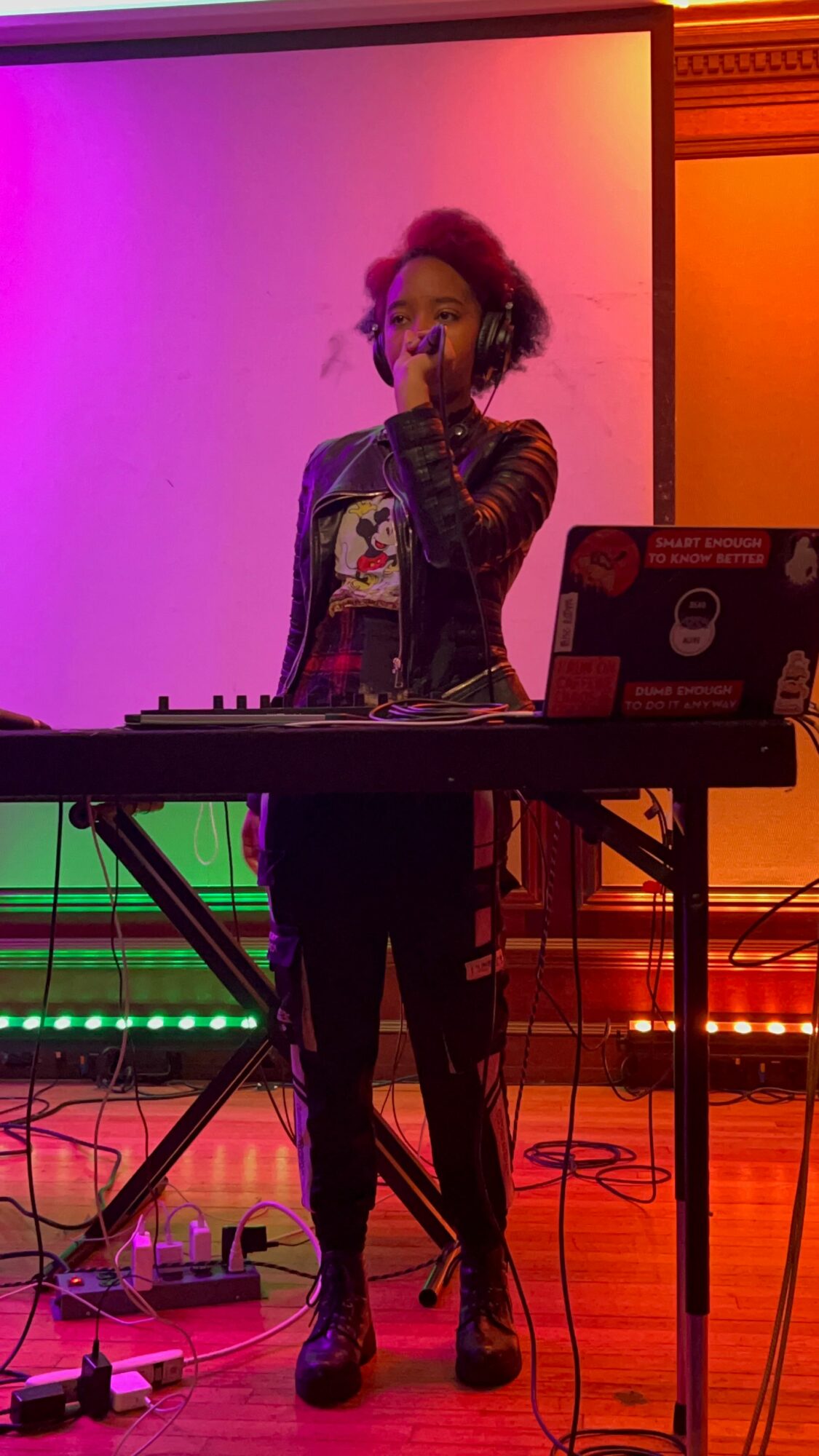Person with curly hair performing with a microphone at a music setup, colorful background lighting, standing behind a keyboard.