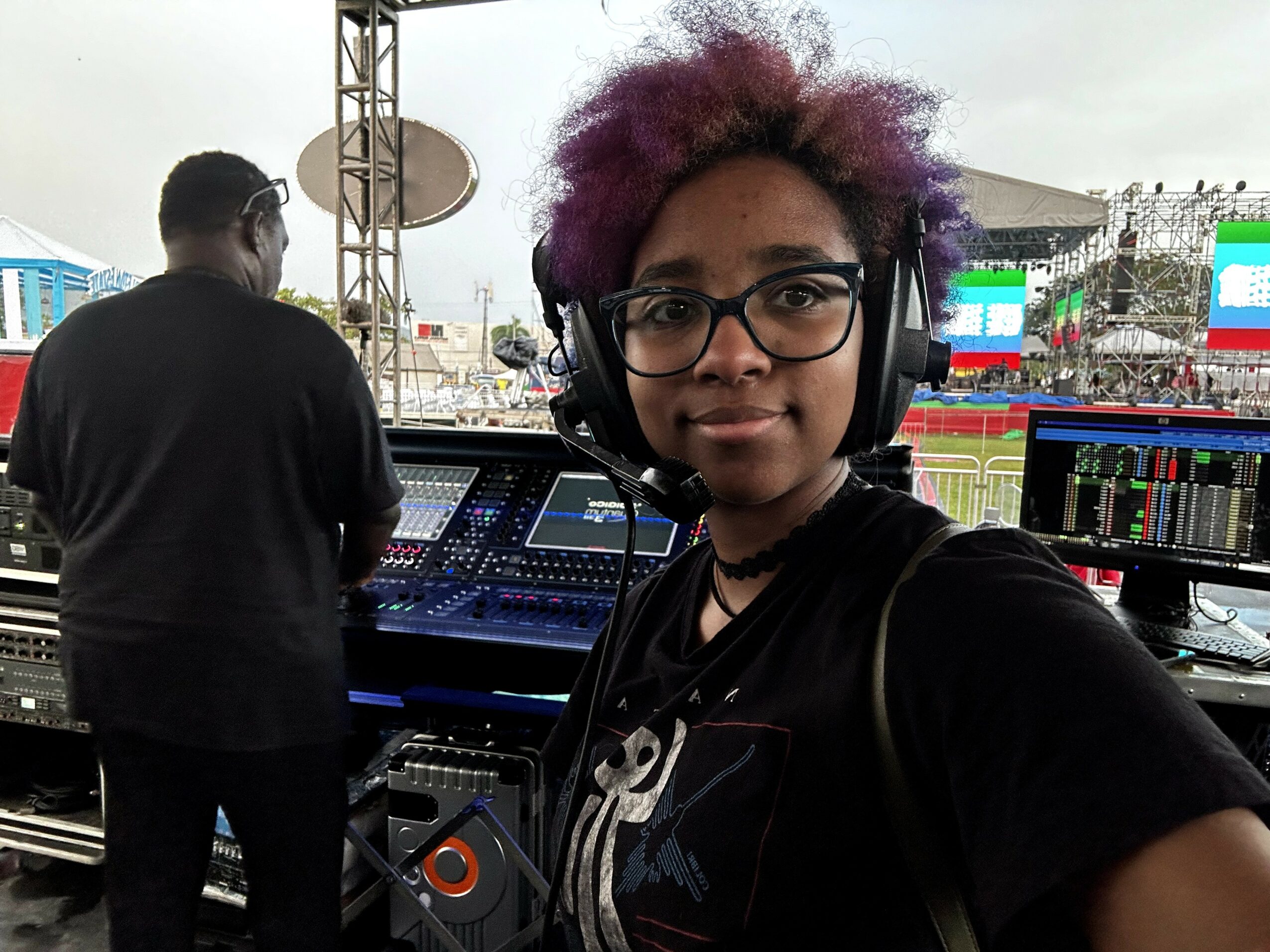 Young woman with purple hair and glasses wearing headphones, operating sound equipment at an outdoor event.