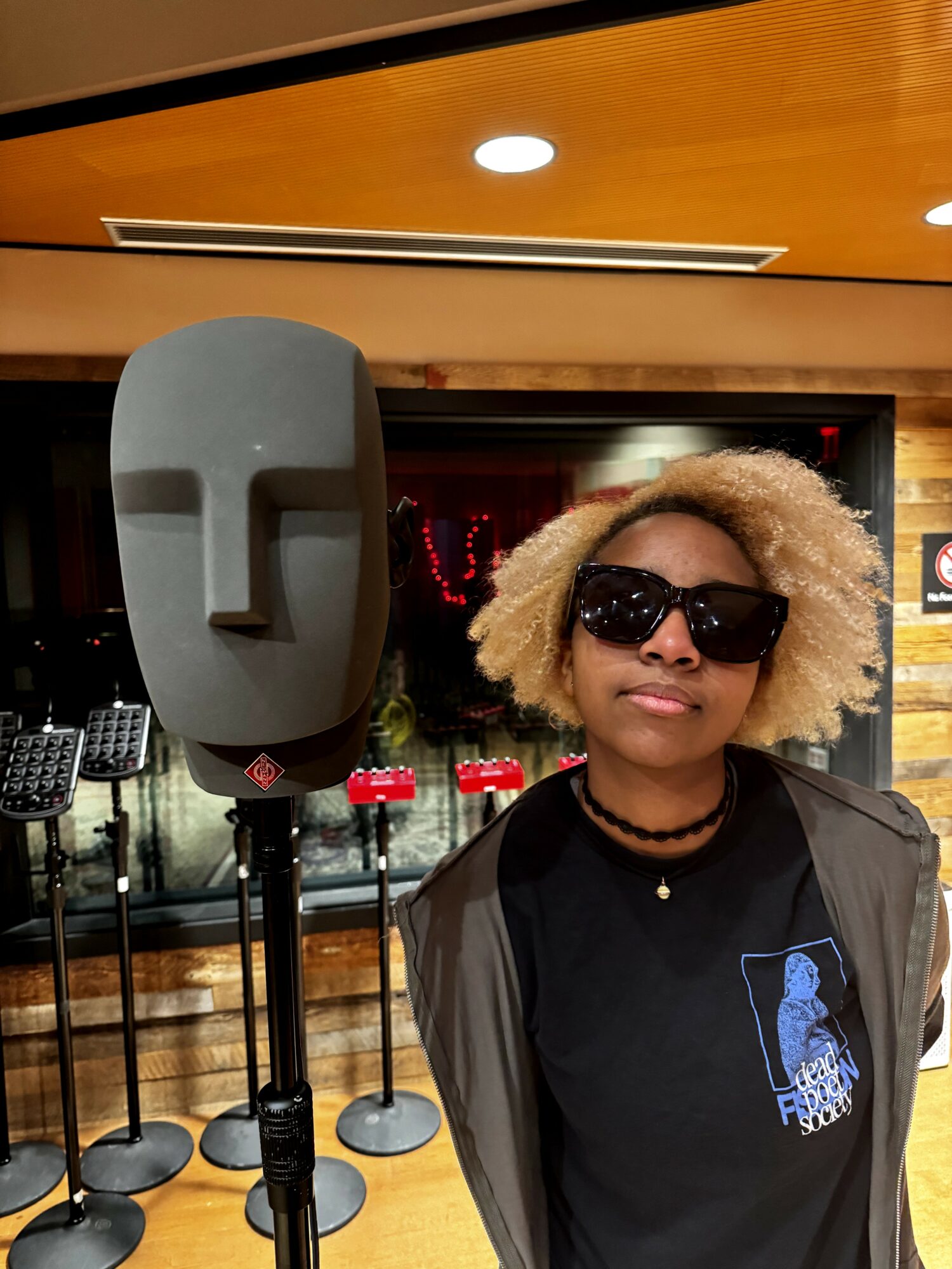 Woman with curly blonde hair wearing sunglasses and a black shirt, standing indoors near a large speaker.