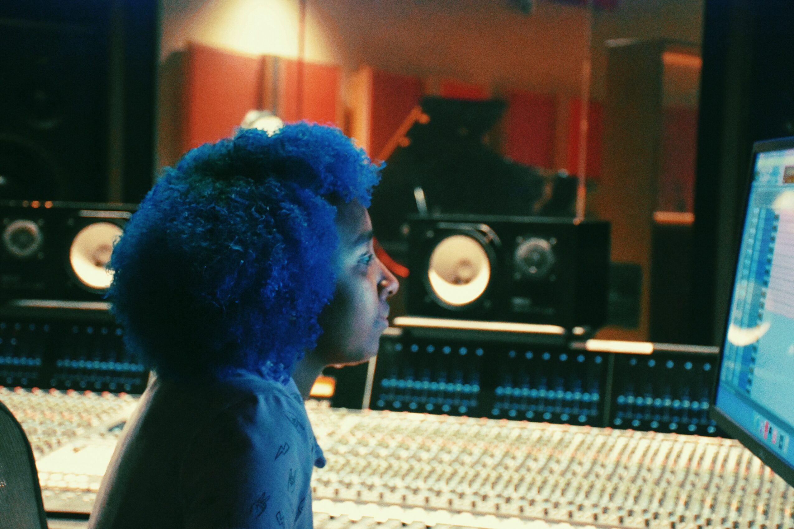 Person with blue hair working at a music mixing console in a studio with speakers and computer screen.